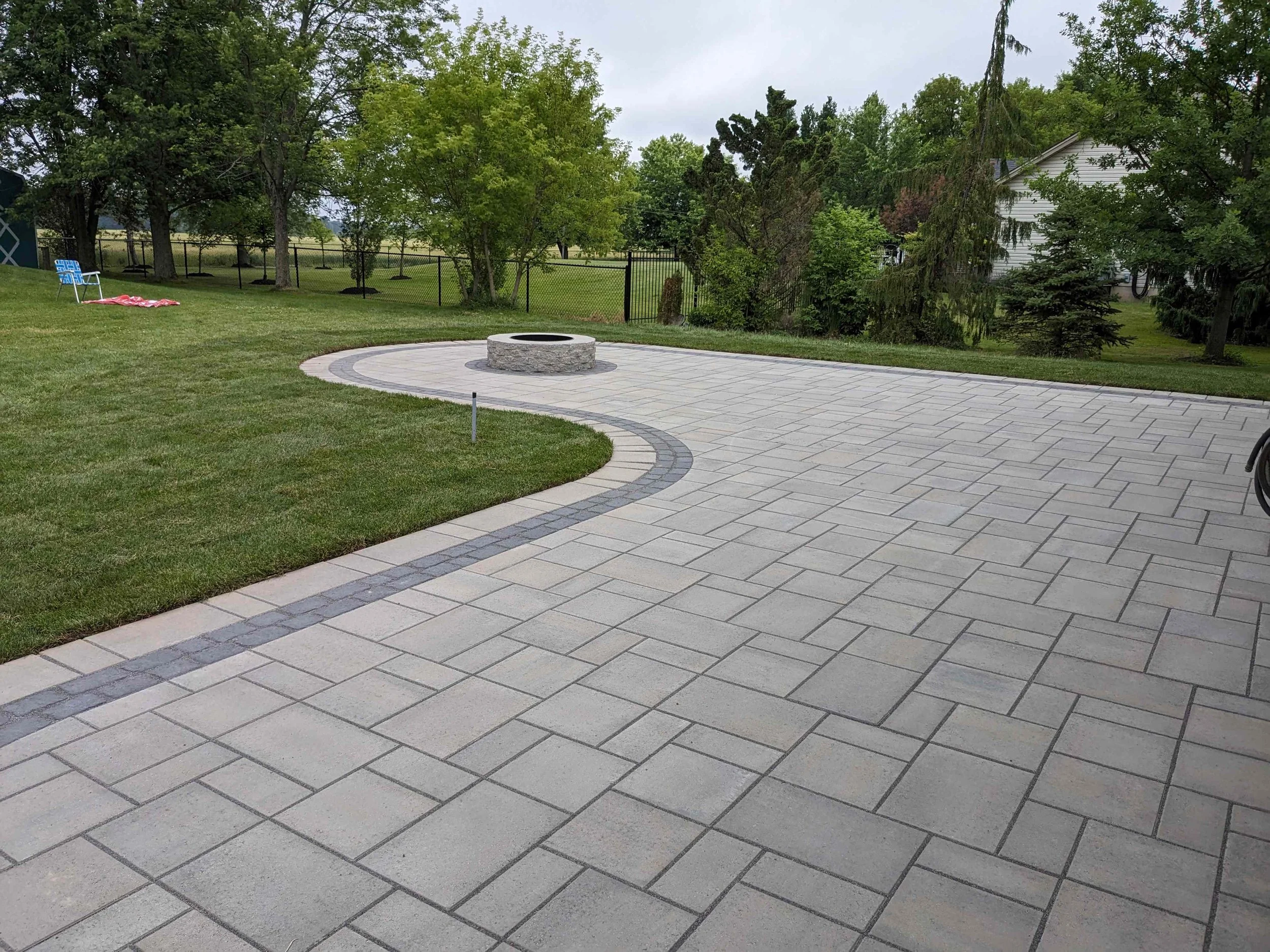 A curved backyard patio featuring Techo-Bloc Blu 60 pavers in Champlain Grey, accented by a onyx black Villagio cobble border and a central stone fire pit