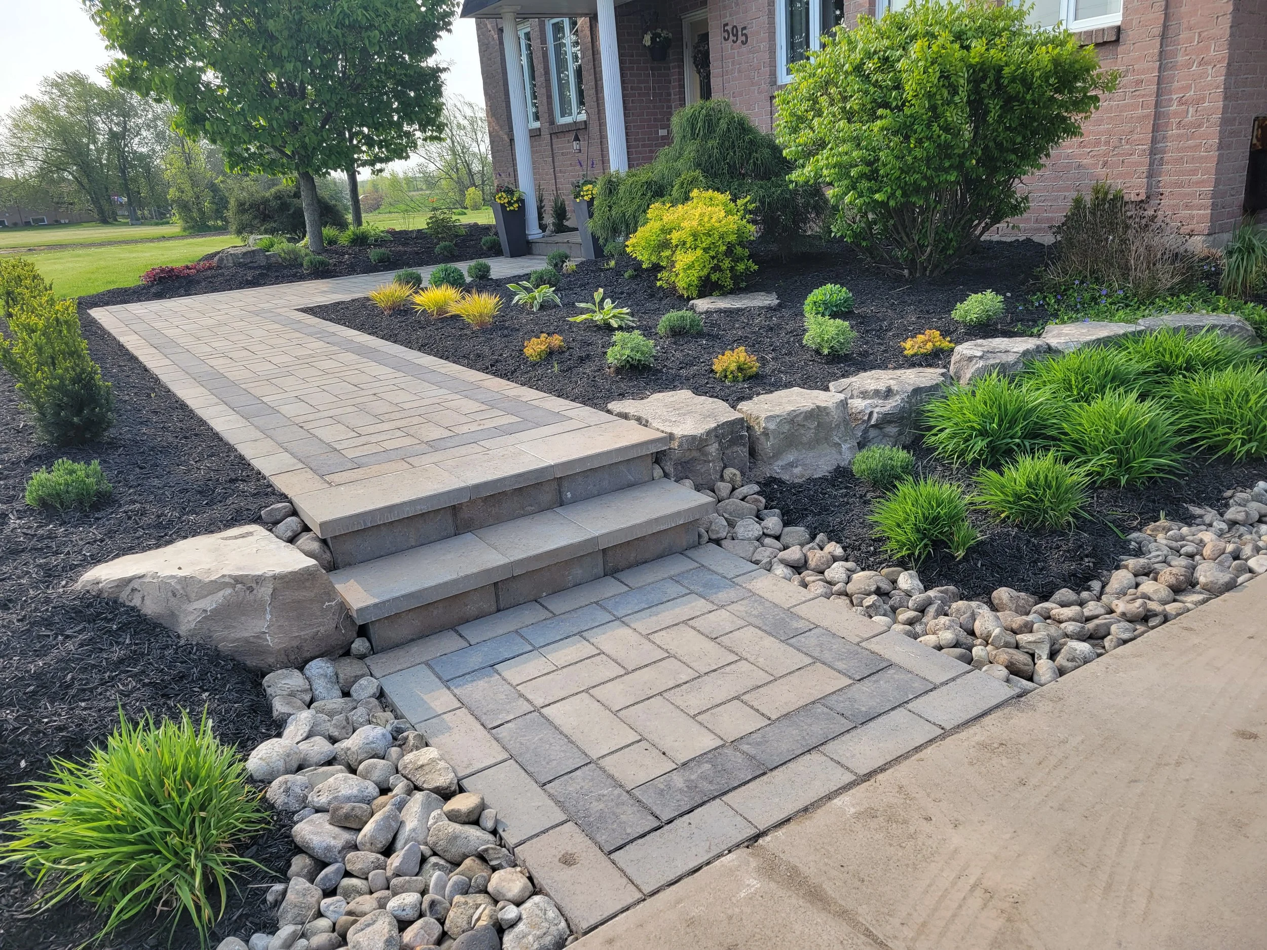 A professionally designed interlock paver walkway with integrated stone steps leads through a landscaped garden bed. The path features Techo-Bloc Blu 60 Pavers, complemented by a natural armor stone retaining wall.