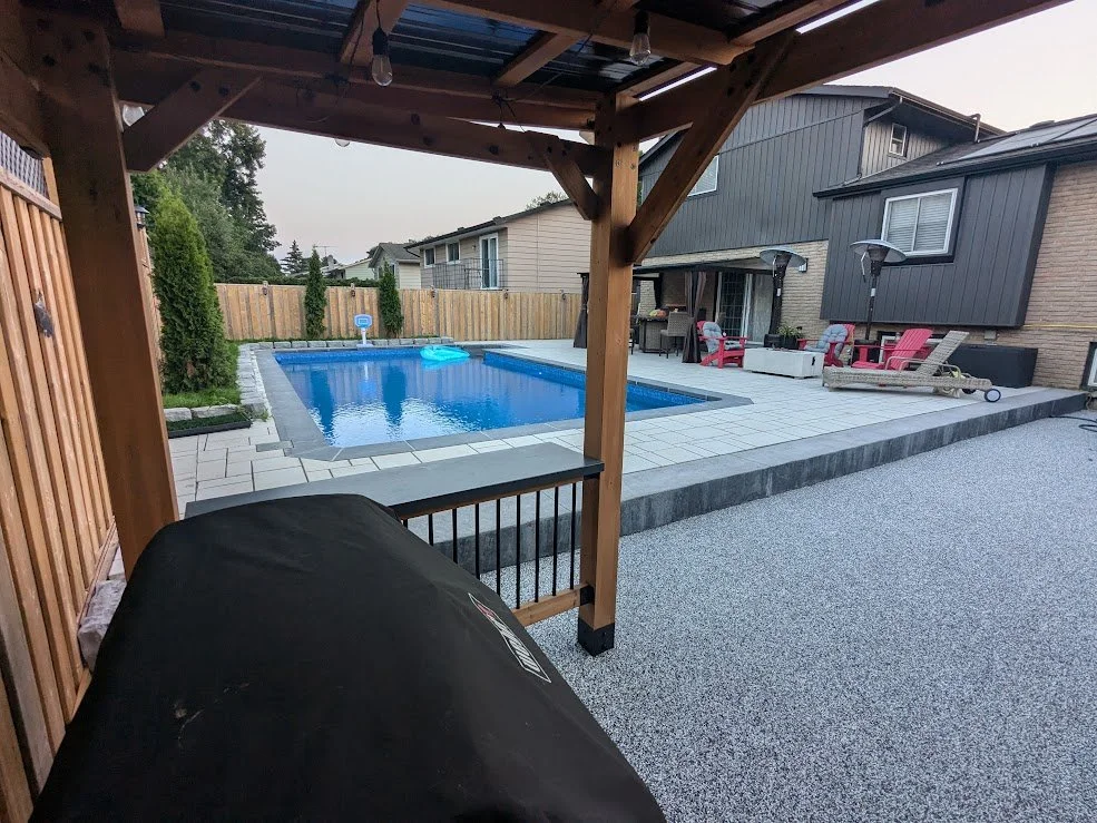 A modern backyard oasis featuring a rectangular inground pool with dark grey coping, a large light-grey paver patio. The space includes a wooden pergola with a built-in bar, and red Adirondack chairs.