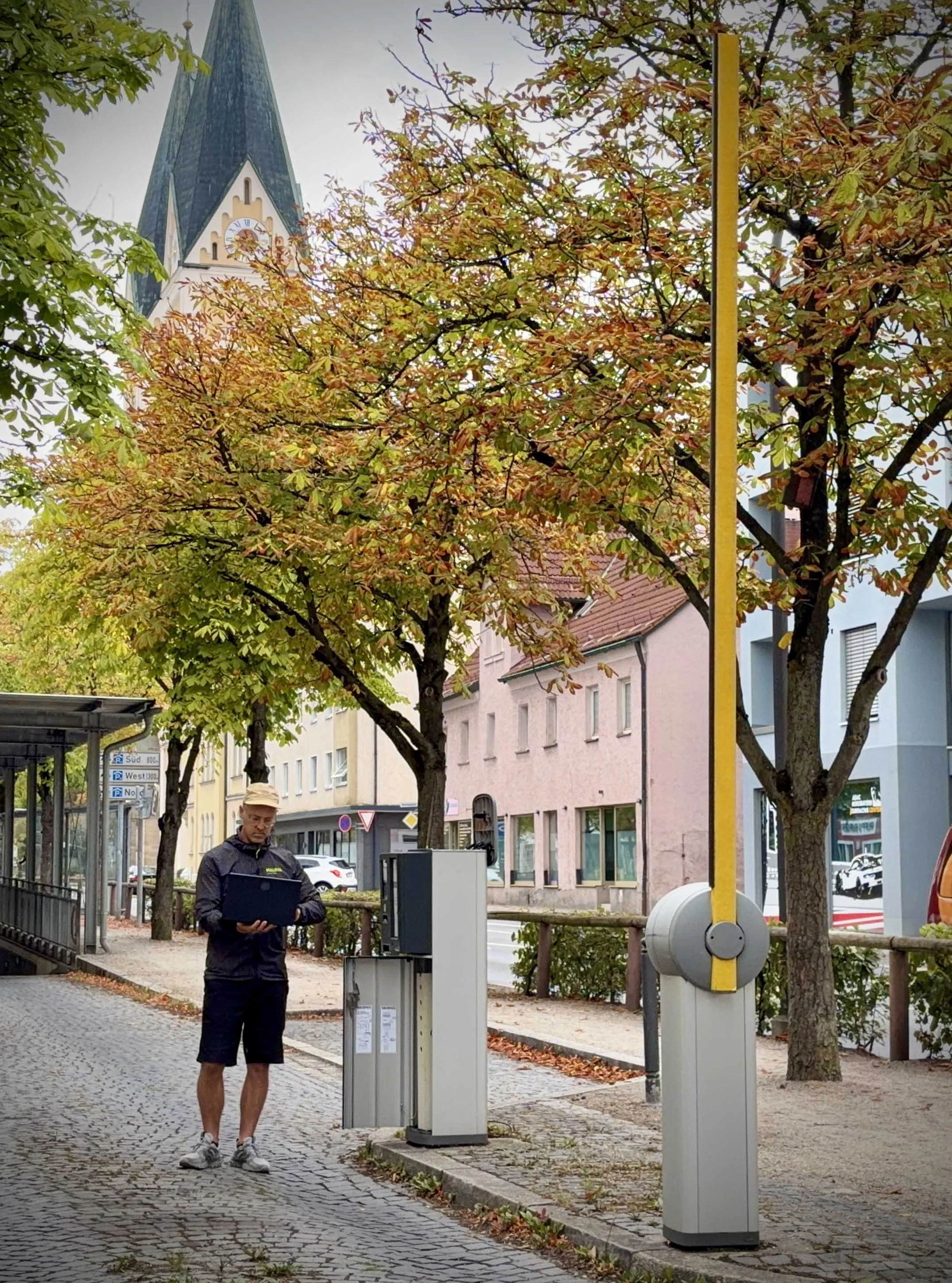 Ein Mann steht mit einem Laptop in einer Fußgängerzone, im Hintergrund sieht man Bäume mit Herbstlaub, ein Gebäude mit Kirchturm und eine Straßenkreuzung. Offener Schranke.
