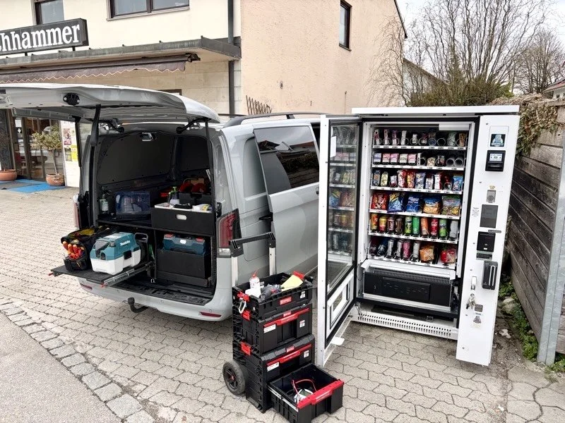 Ein Lieferwagen mit geöffneten Rück- und Seitentüren, der mit Werkzeugen und Batterien ausgestattet ist, sowie einer Snack- und Getränkeautomat auf der rechten Seite, der verschiedene Getränke und Snacks enthält.