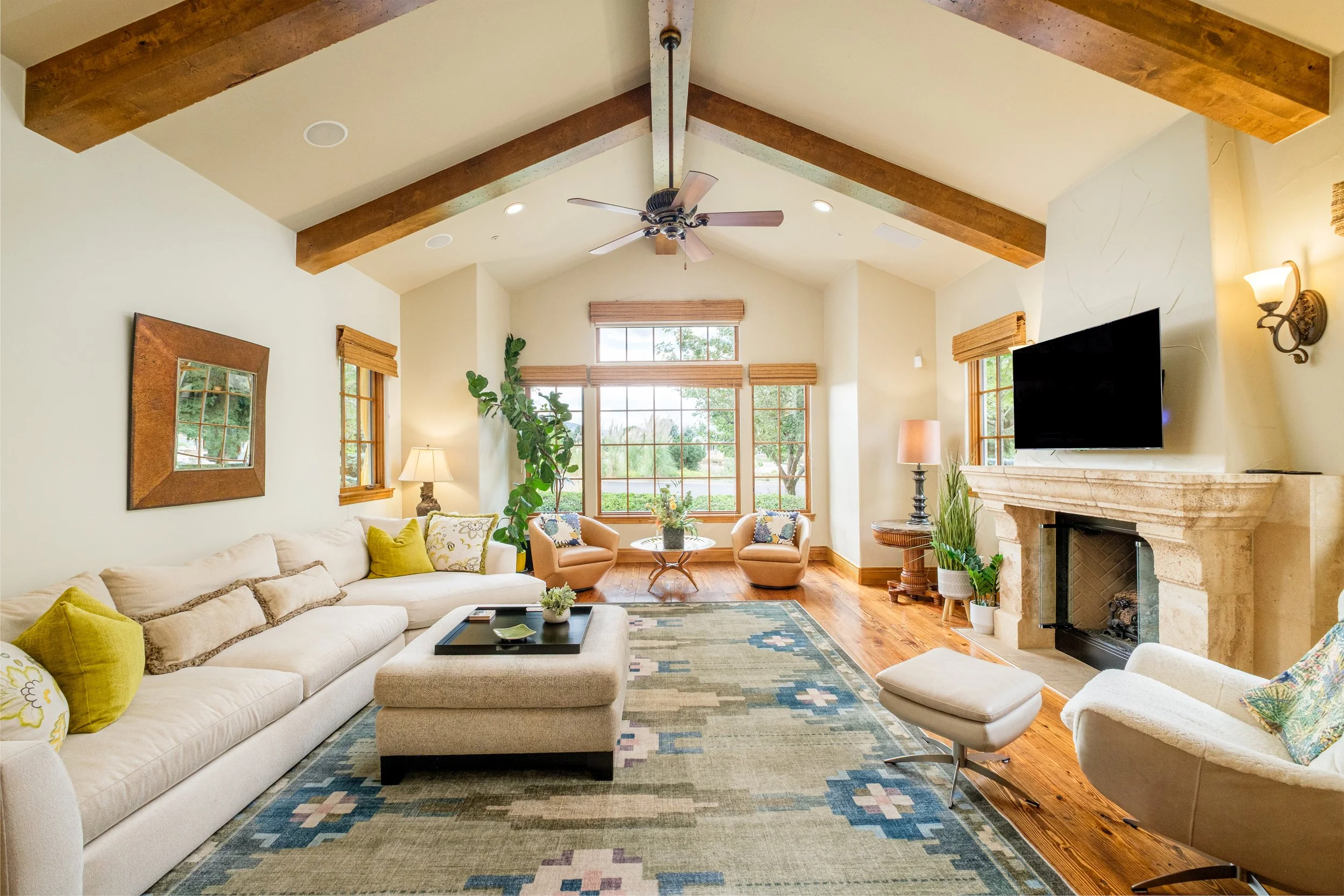A spacious living room with cream-colored walls and a vaulted ceiling with wooden beams. It features a large beige sectional sofa with yellow and floral throw pillows, a rectangular ottoman, a patterned area rug, a fireplace with a television mounted above, large windows with wooden blinds, and various chairs and lamps, creating a cozy and inviting atmosphere.