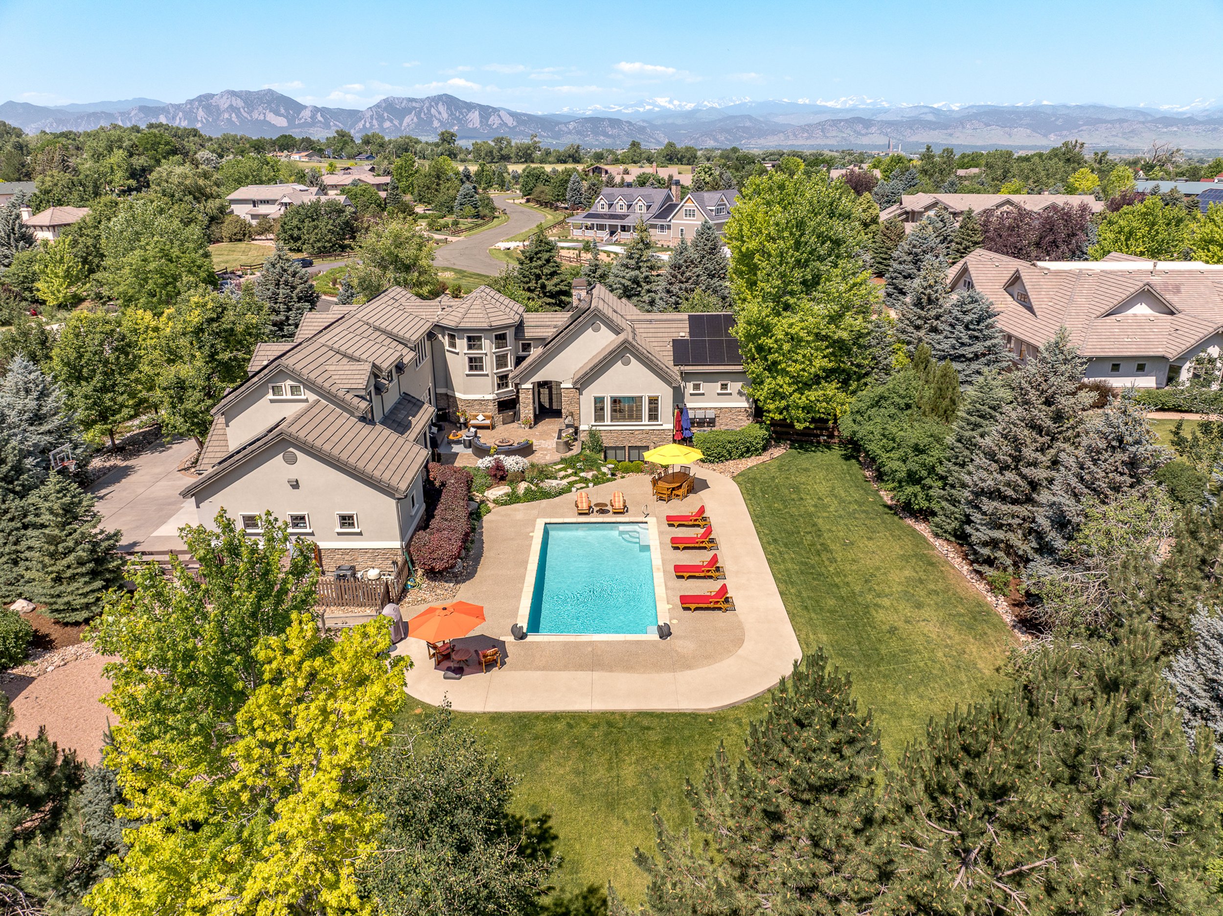 A residential backyard with a swimming pool, lounge chairs, and umbrellas, surrounded by lush green trees, with a neighborhood and mountains in the background.