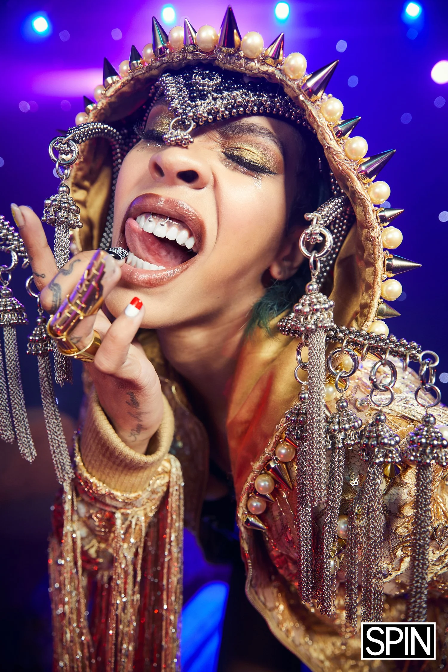 A person wearing a heavily embellished gold hood with spikes and pearls, gold eyeshadow, and jewelry, sticking out their tongue while holding up their hand with rings in a lively, colorful setting.