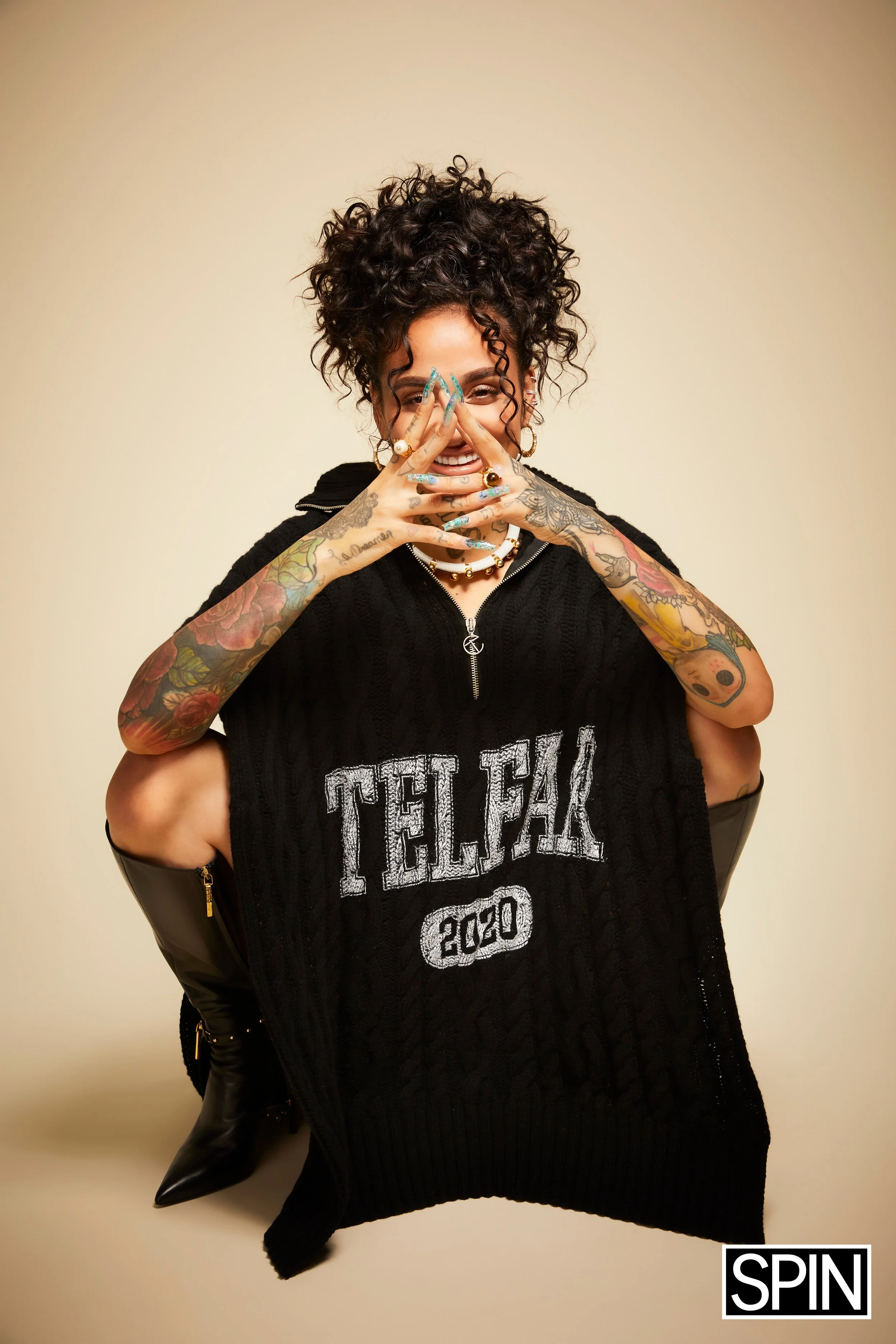 A woman with curly hair and tattoos is smiling and covering her face with her hands, wearing a black sleeveless vest with 'TELL FAK 2020' written on it, and sitting against a beige background.