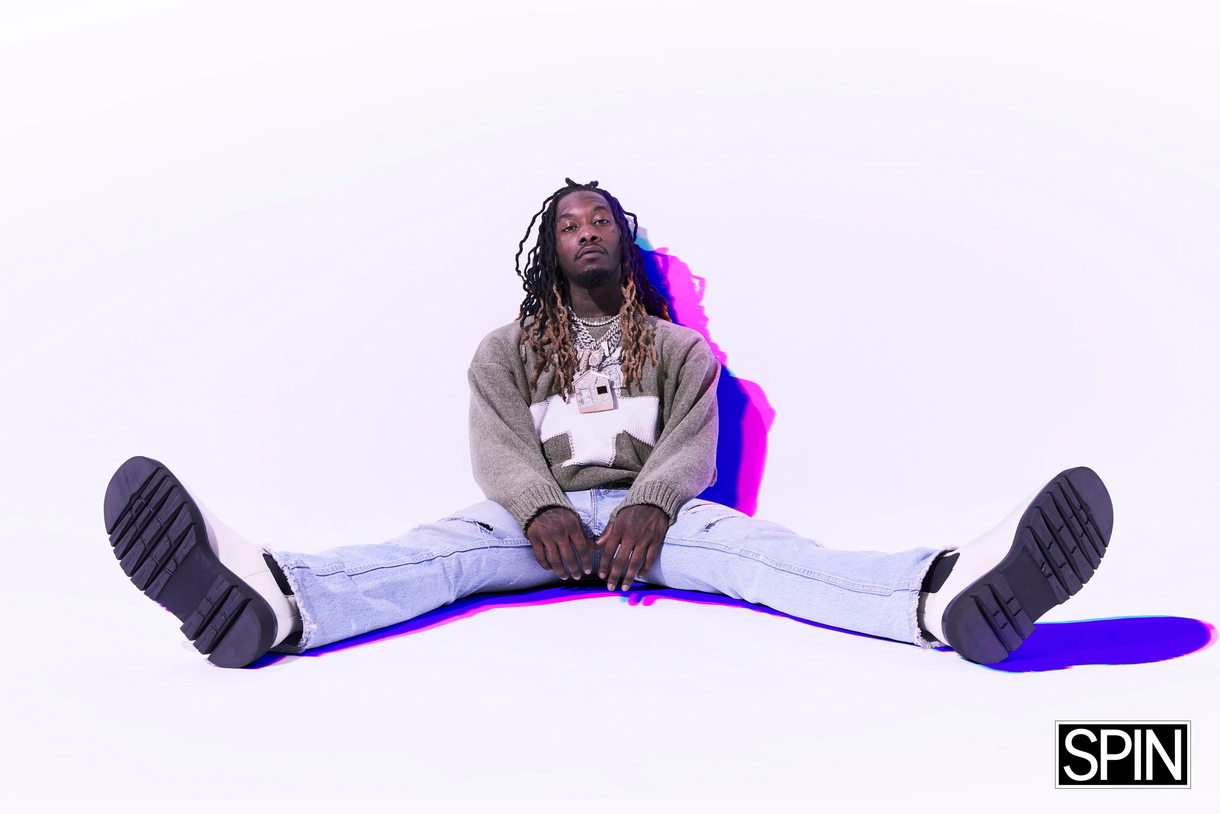Man sitting on the floor with legs spread wide apart, wearing a sweater, ripped jeans, and sneakers, against a white background with a colorful purple and pink shadow, and the word 'SPIN' in the bottom right corner.