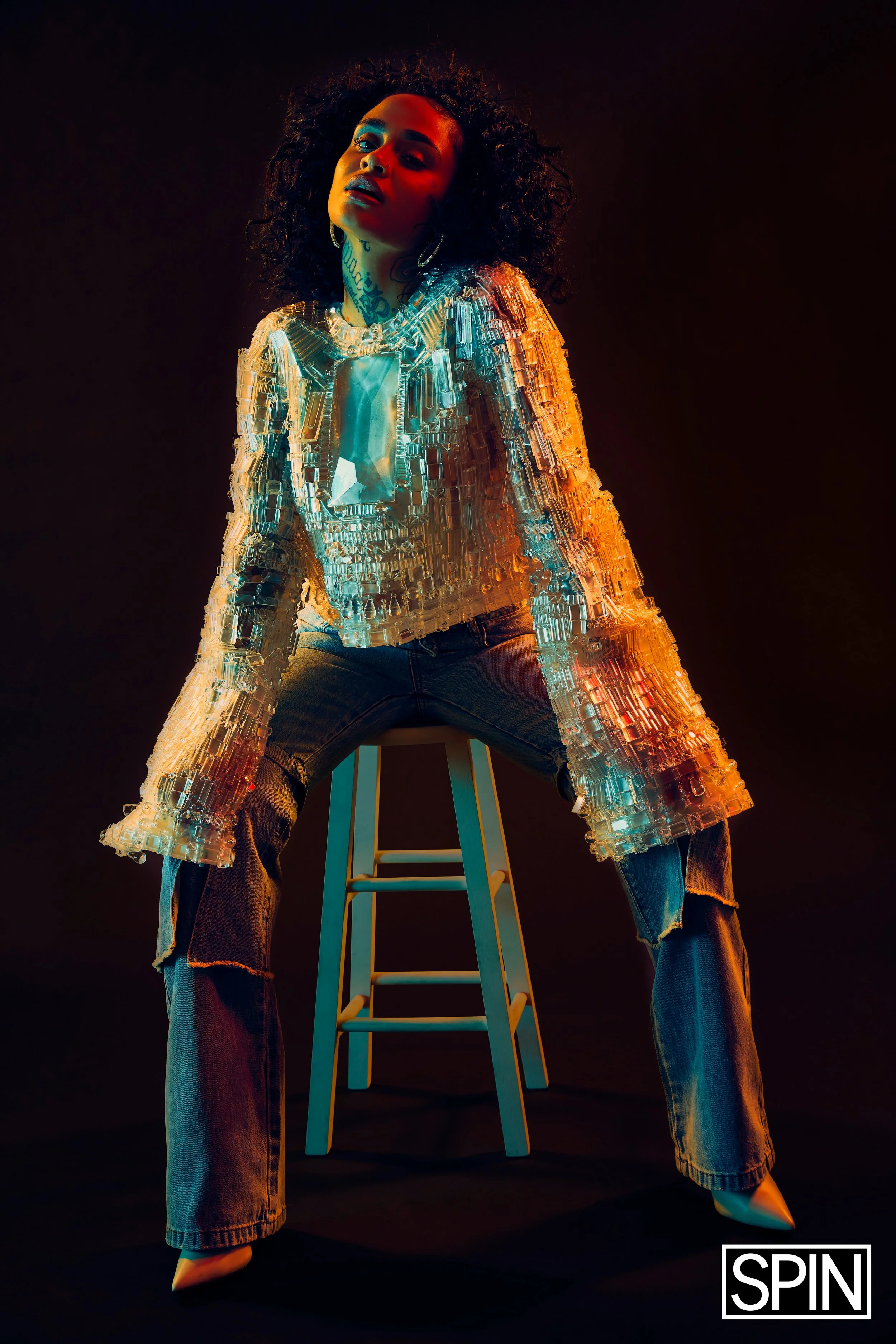 A woman with curly hair sitting on a blue stool against a dark background, wearing a shiny, reflective, crystal-like jacket, dark jeans, and high-heeled shoes, with colorful lighting highlighting her features.