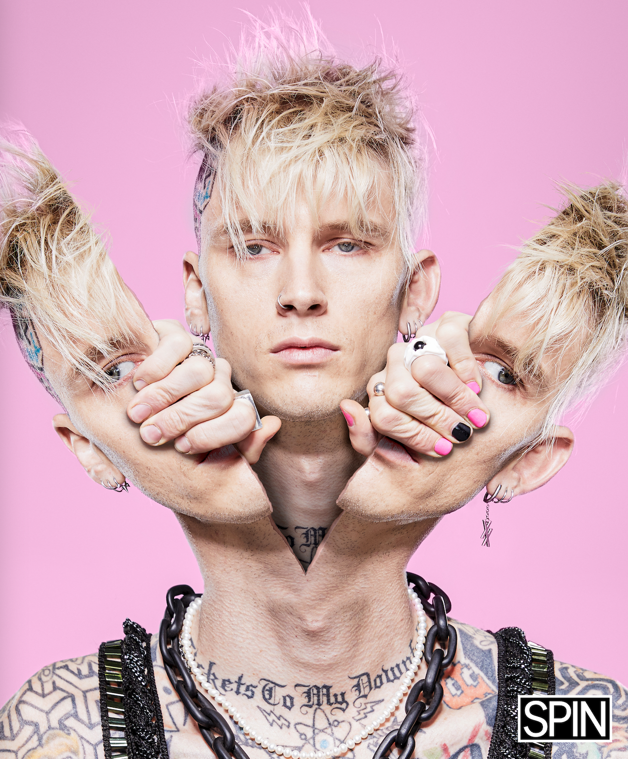 A person with blond, spiky hair has their face edited to show two additional faces on each side, held by hands with pink and black nail polish, against a pink background. The person has tattoos, piercings, and is wearing jewelry, including necklaces 