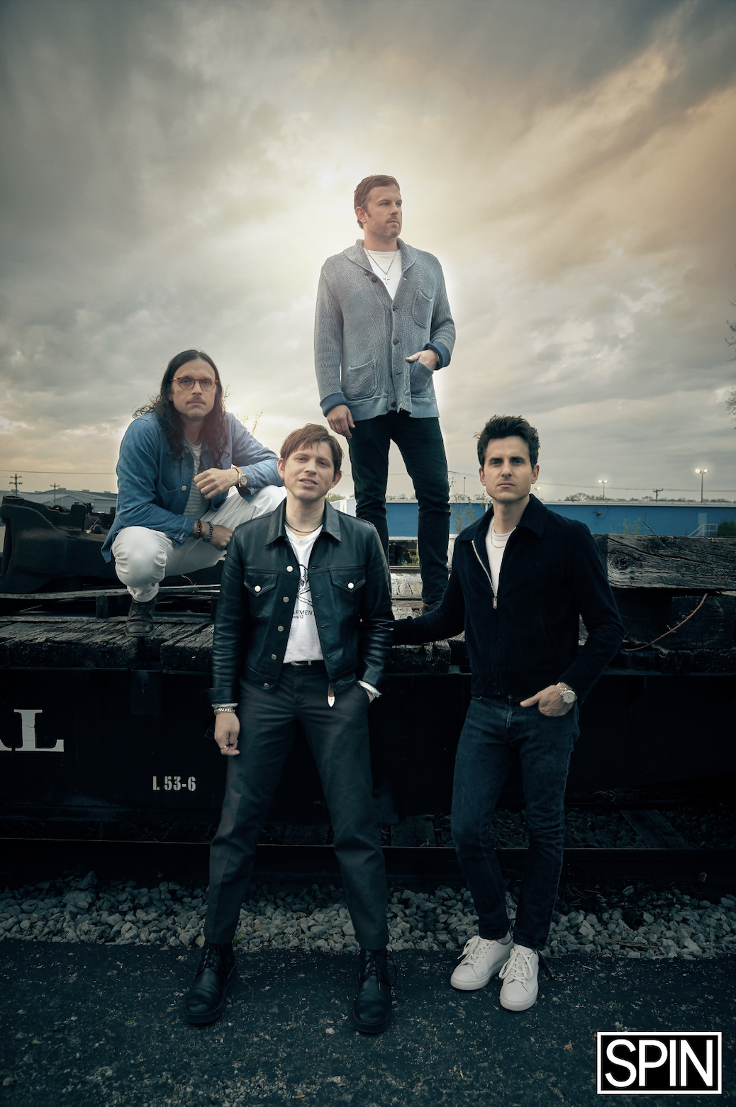 Four men posing on railroad tracks under a cloudy sky, with one man standing on a railcar and the other three standing or kneeling nearby. The photo has a moody, artistic feel.