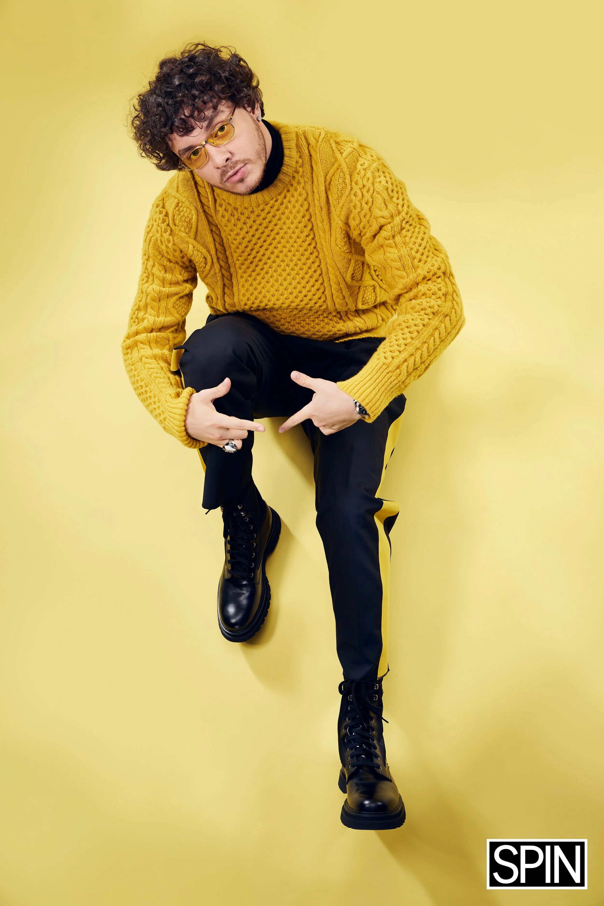 A person with curly hair, glasses, wearing a yellow sweater and black pants with yellow accents, squat on a yellow background. They are wearing black combat boots and accessorized with rings and a watch. The bottom right corner has the SPIN logo.