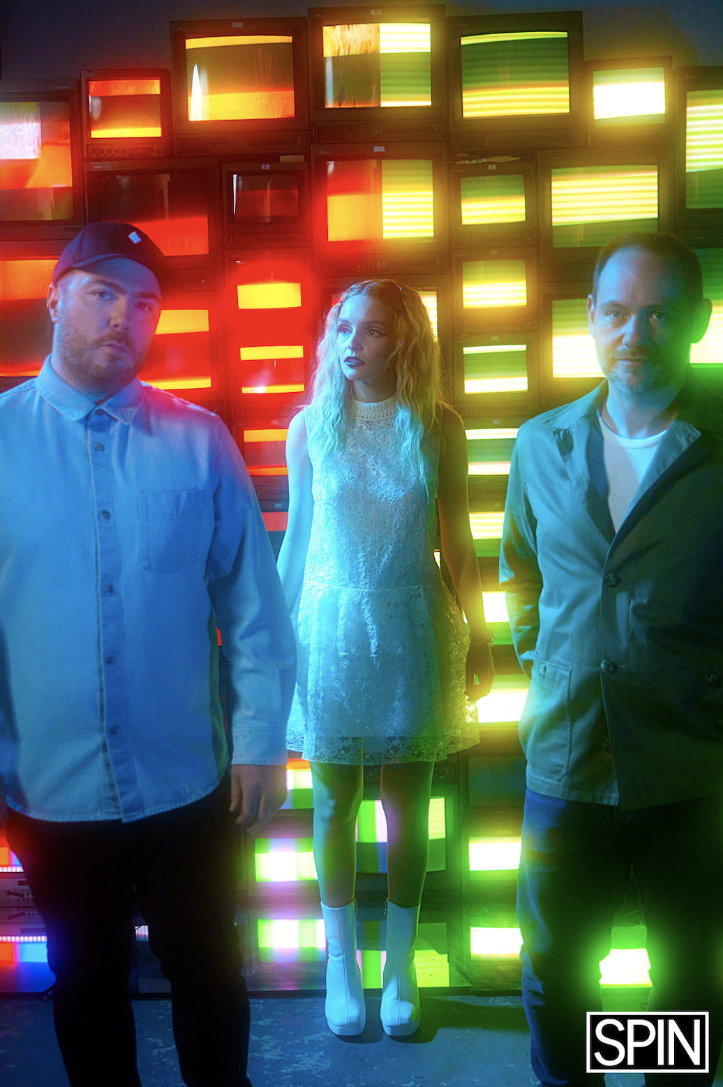 Three people standing in front of a colorful, illuminated backdrop of retro televisions emitting red, yellow, and green lights.