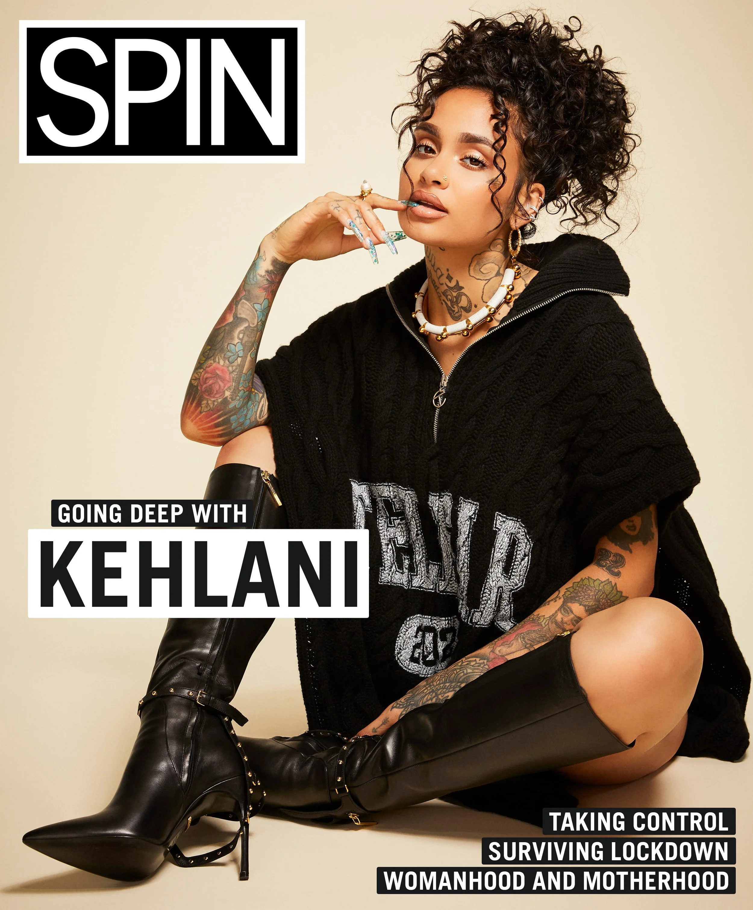 Kehani sitting on the floor in black boots and a oversized black hoodie, with tattoos, jewelry, and curly hair, in a portrait style photo.