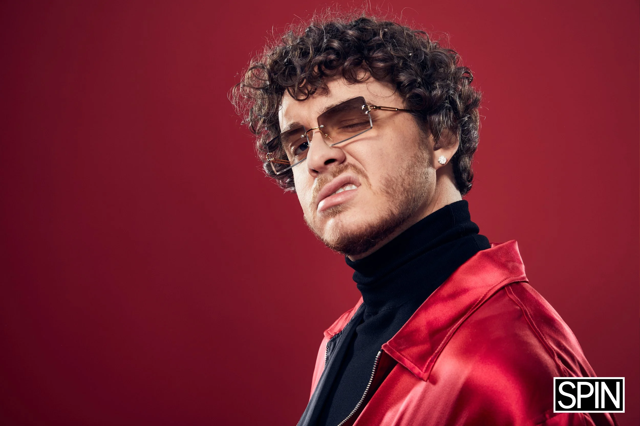 A young man with curly hair, wearing sunglasses, a black turtleneck, and a red satin jacket, standing against a red background, making a grimacing facial expression.