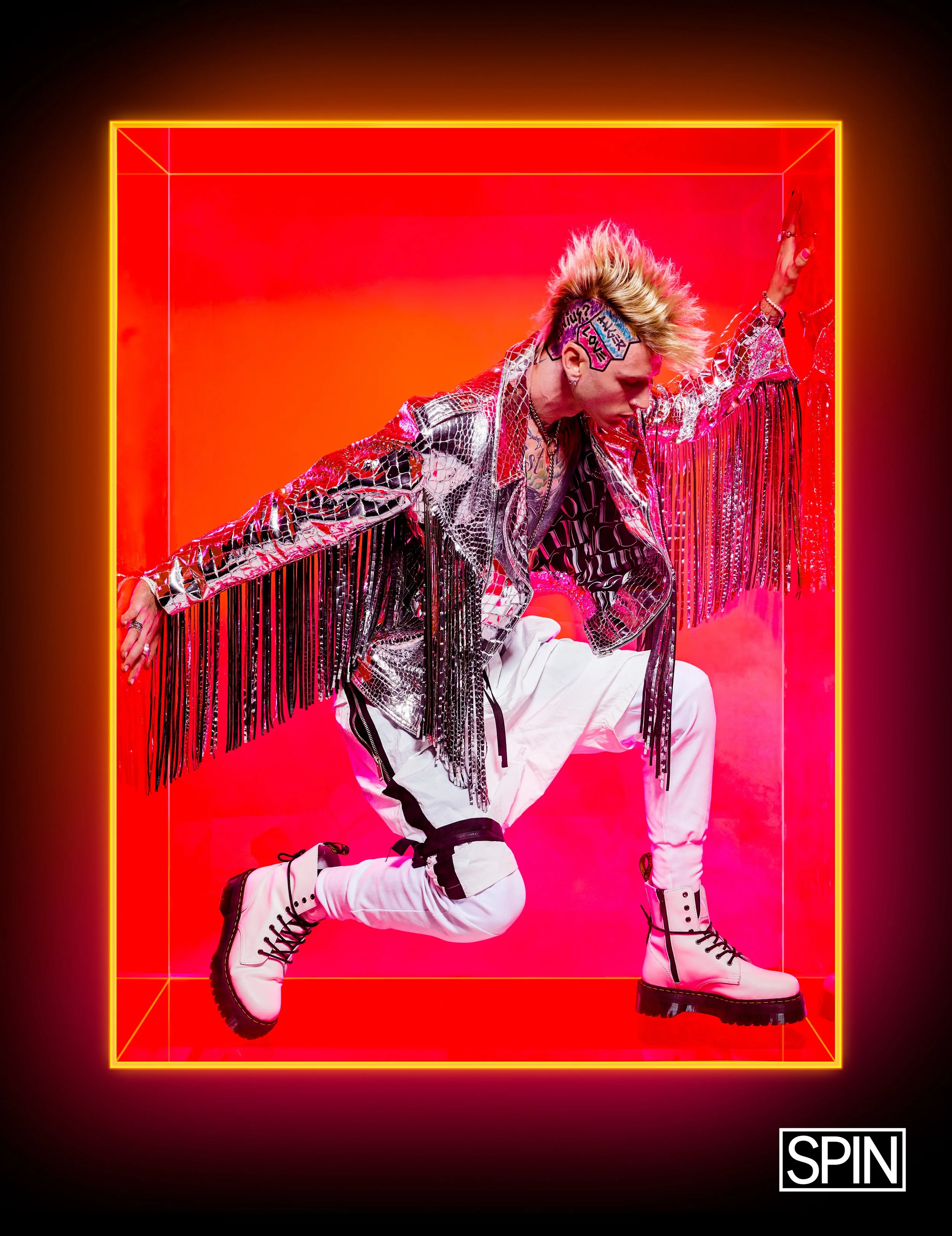 A person with platinum blonde hair styled in a spiky mohawk posing inside a neon-lit box with a red background, wearing a shiny silver and black jacket with fringe, white pants with black accents, and white combat boots.