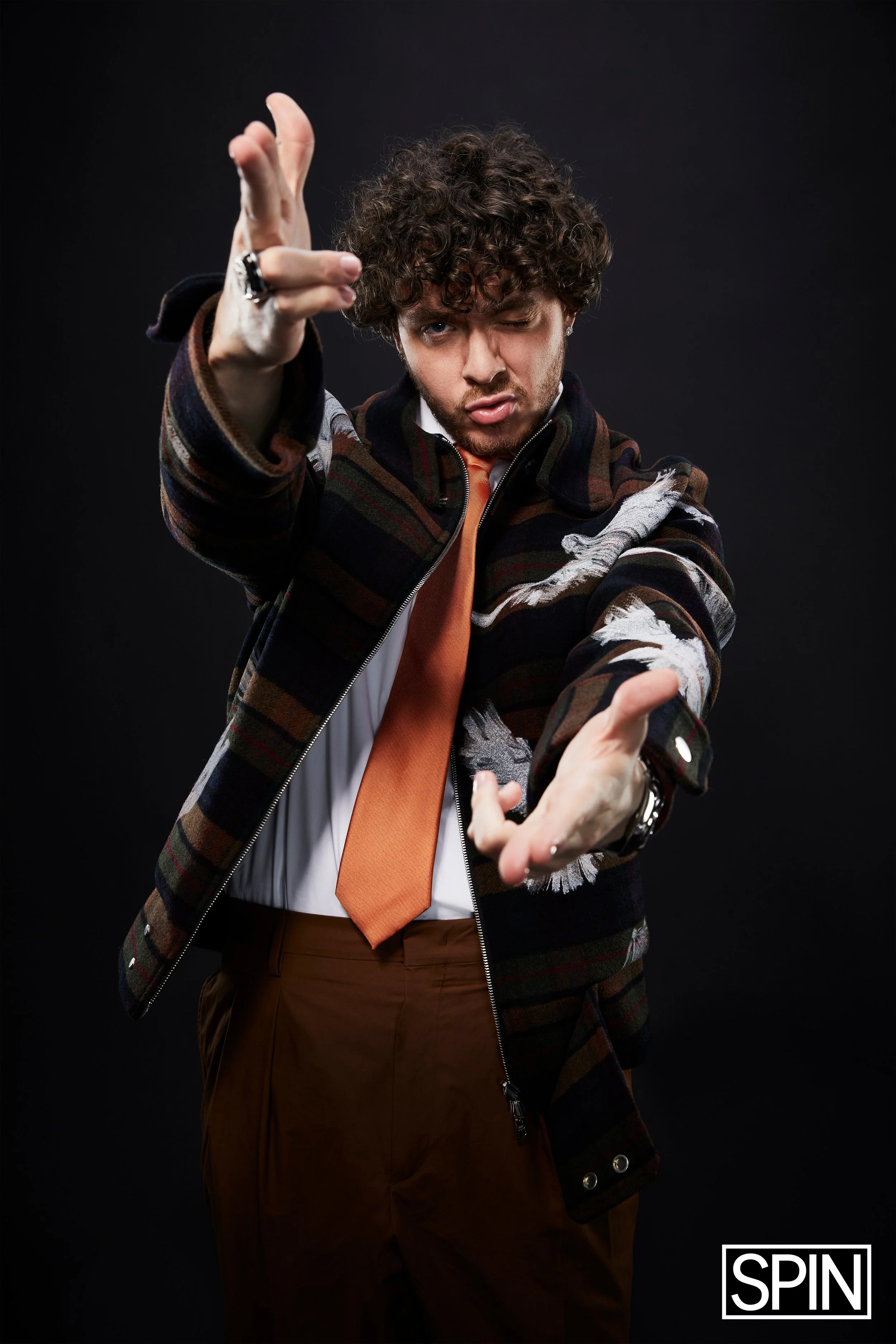 A man with curly hair and a playful expression is posing with his hands extended toward the camera. He is wearing a patterned jacket, a white shirt, an orange tie, and brown pants on a black background with a logo that says 'SPIN' in the bottom right