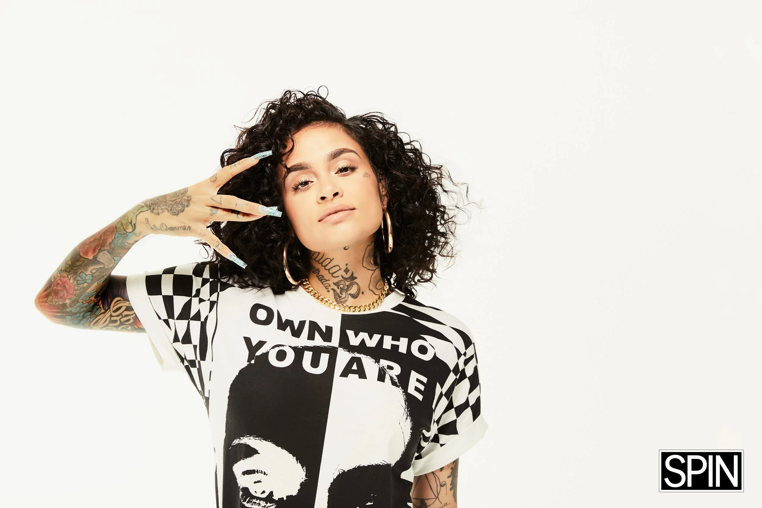 A woman with curly black hair and tattoos on her arms and neck, wearing a black and white graphic t-shirt with the text 'OWN WHO YOU ARE' and an image of a woman's face. She is posing with her hand near her face against a plain white background, and 