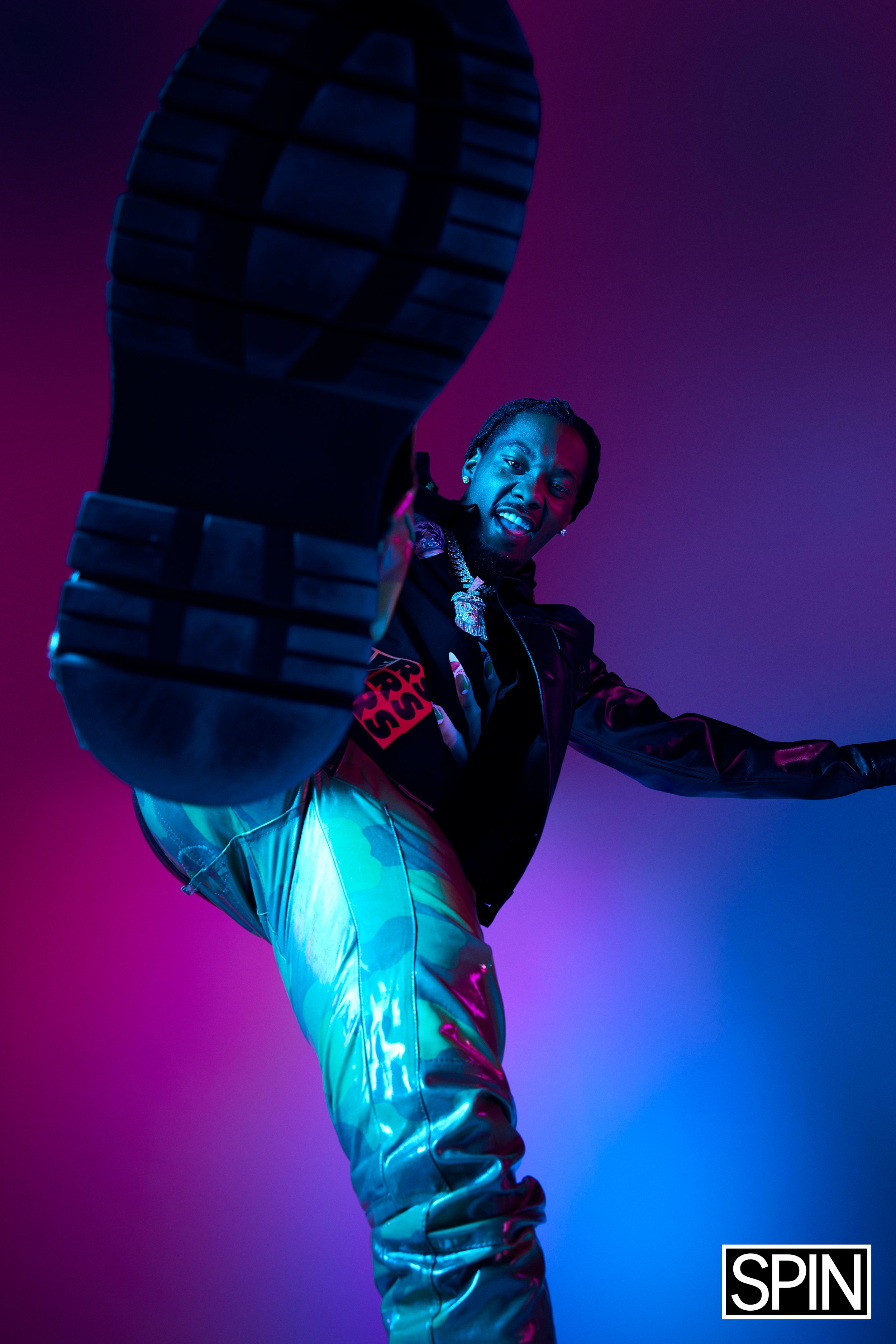 A man with dreadlocks and a beard is Minnesotan in the air, looking down at the camera with a smile, against a colorful purple and blue background. The man is wearing a black jacket, shiny metallic pants, and jewelry.