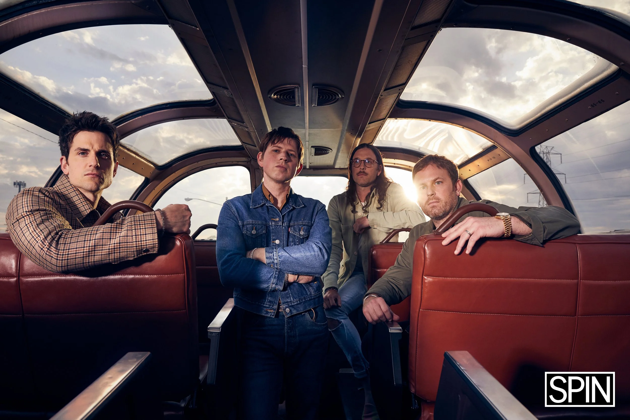 Four men with serious expressions inside a bus with large windows, showing a cloudy sky outside, the sun shining through the window, and a SPIN logo at the bottom right corner.