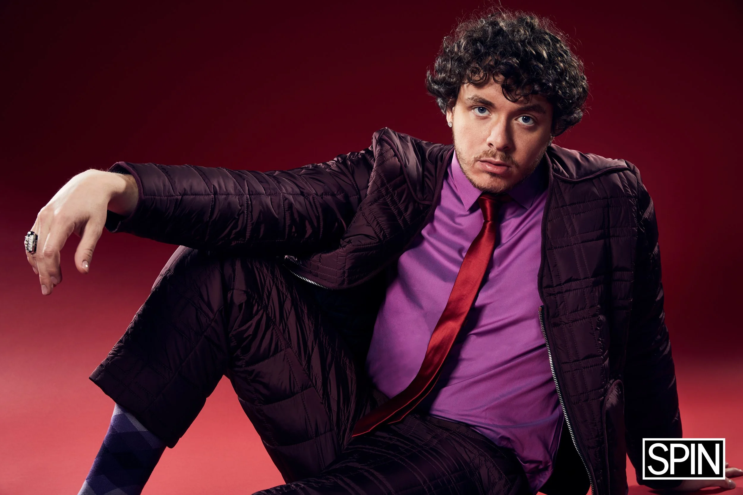 A young man with curly hair poses against a red background, wearing a dark purple quilted jacket, a purple shirt, a red tie, and patterned pants. The SPIN logo appears at the bottom right corner.