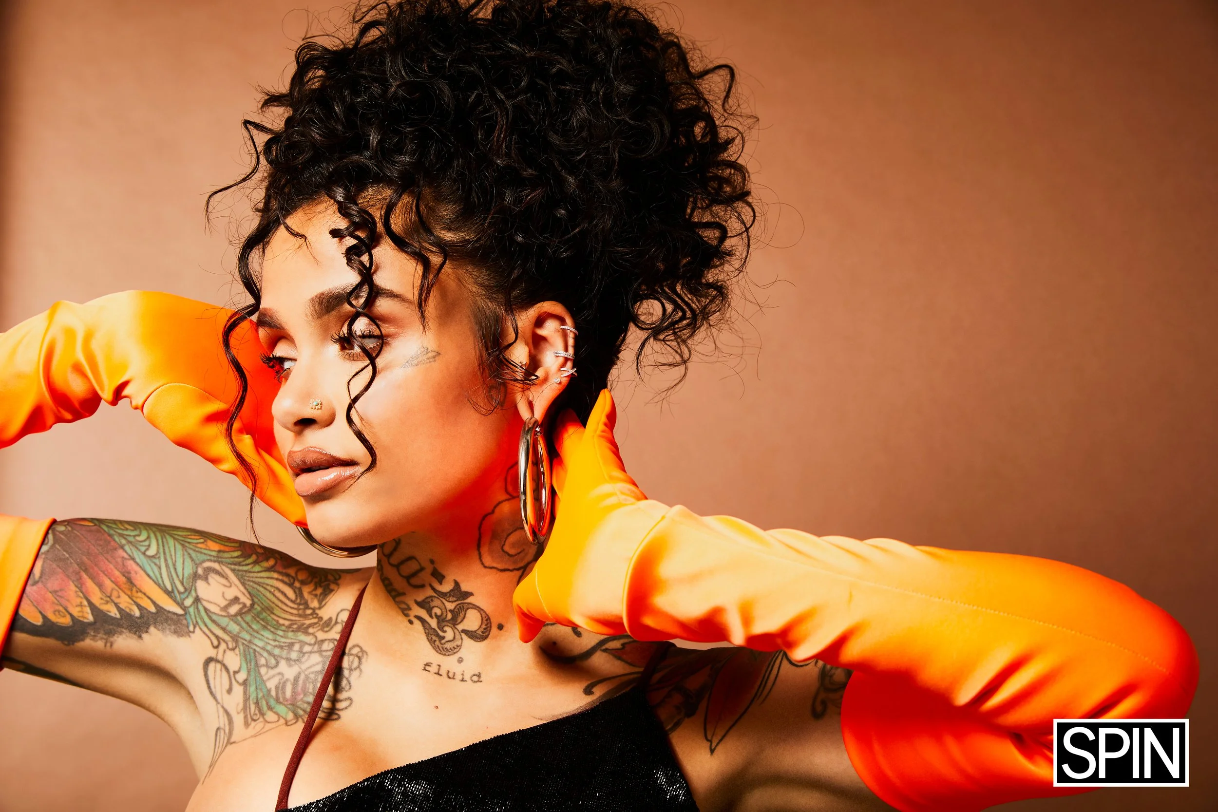 A woman with curly black hair, tattoos on her arms and chest, wearing large hoop earrings, a black top, and orange satin gloves, poses against a plain background.