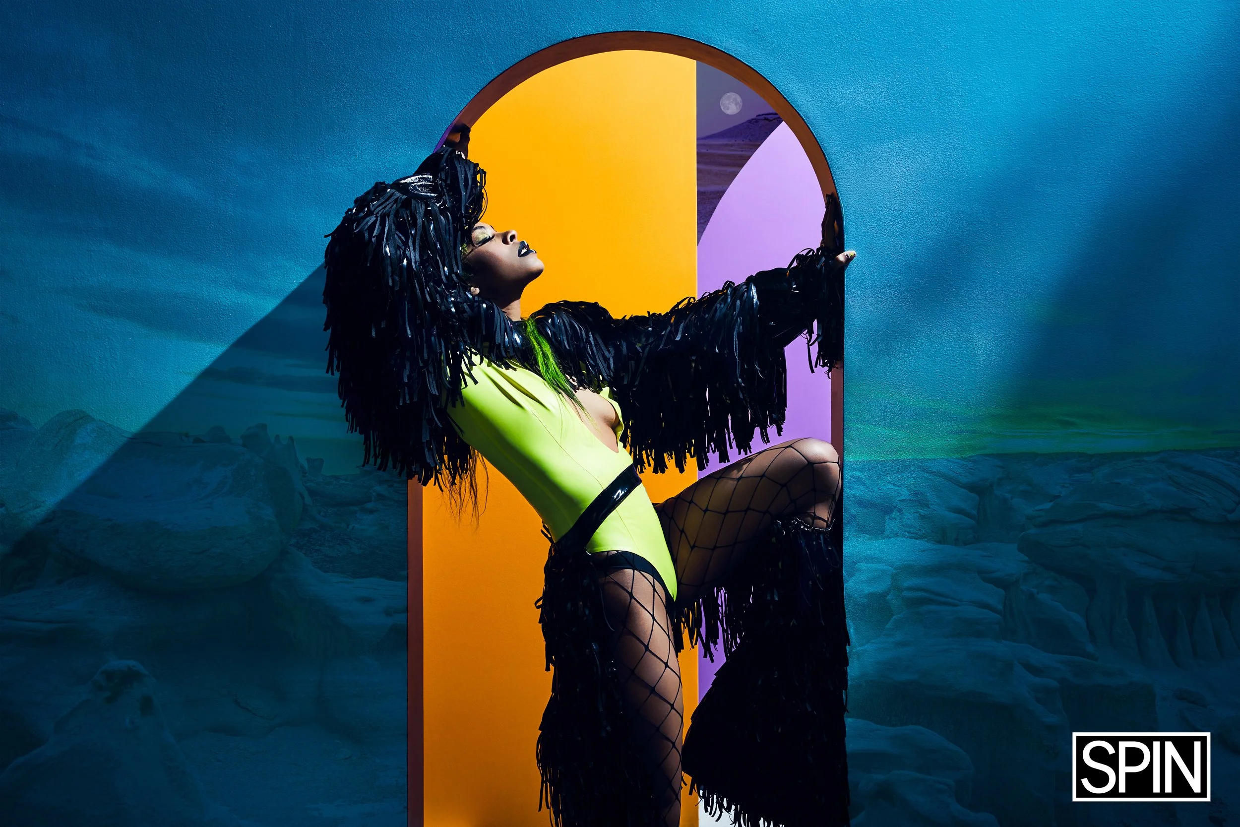 A woman with dark skin and long black yarn-like hair is posing in front of a colorful background, wearing a bright yellow bodysuit, fishnet stockings, and a black fringed jacket, with her eyes closed and head tilted back.