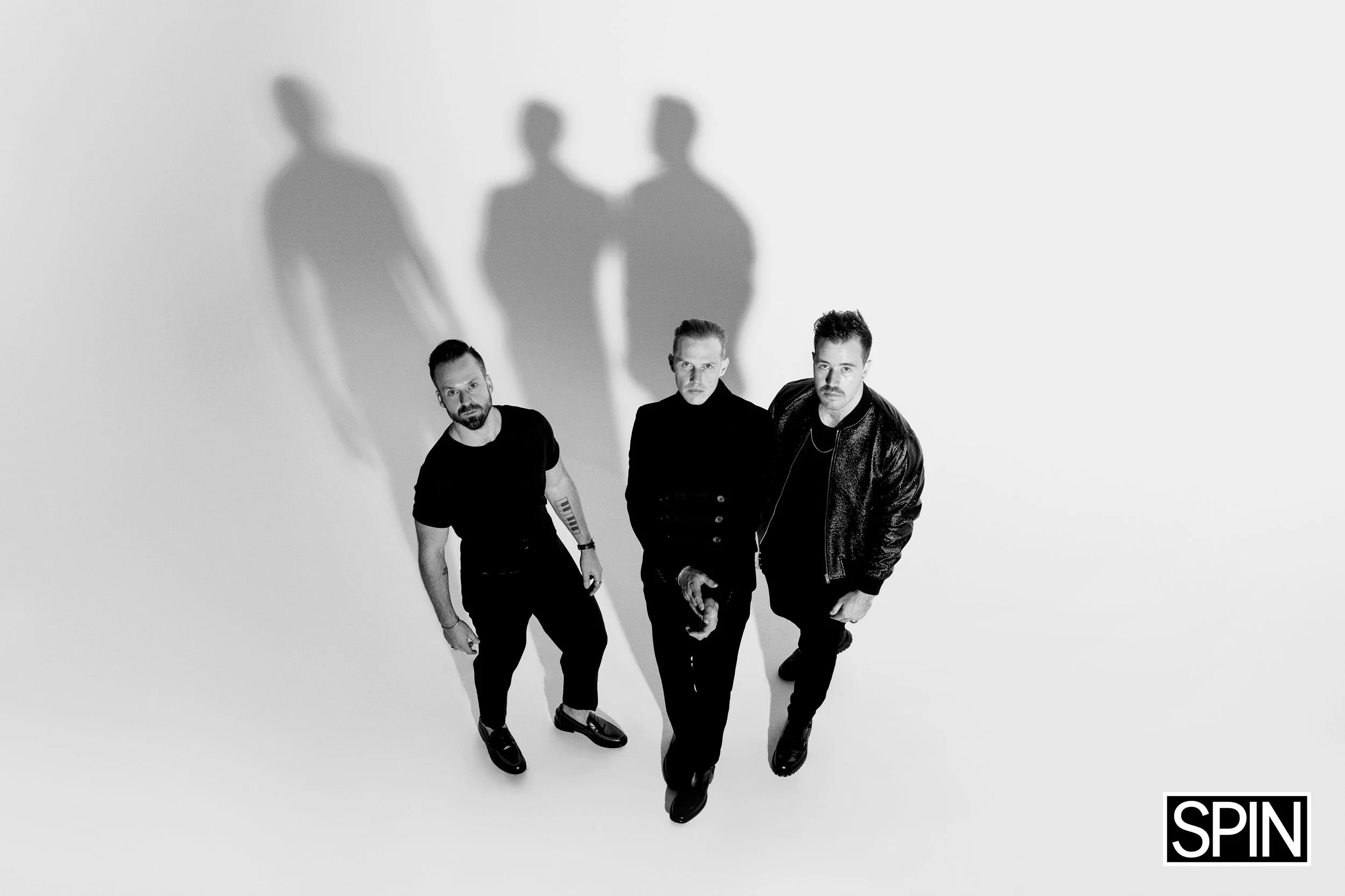 Black and white photo of three men walking toward the camera from a high angle, with their shadows projected on the wall behind them. The man on the left has a beard and tattoo on his arm, wearing a black t-shirt and dark pants. The man in the middle
