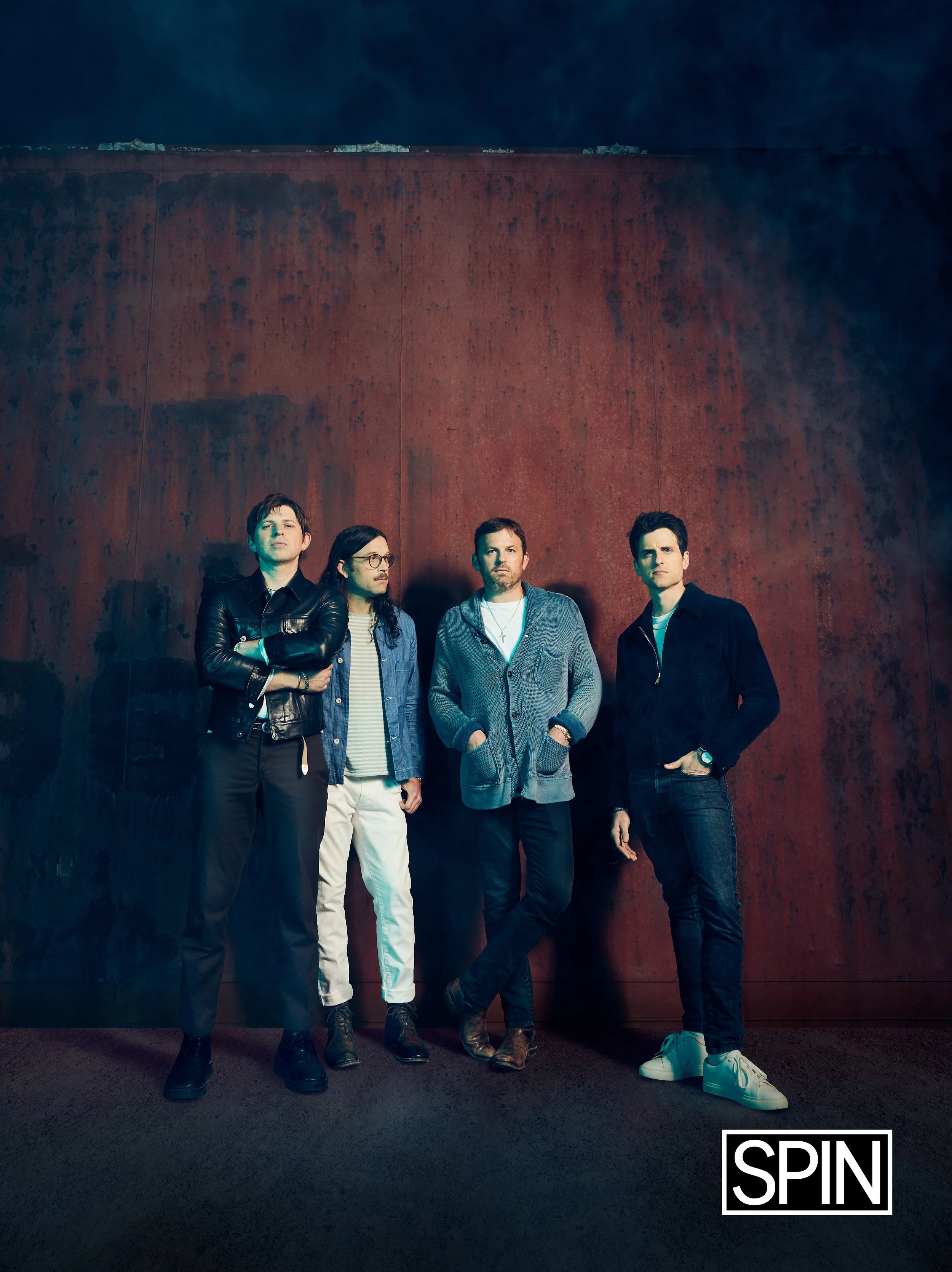A group of four men standing against a rust-colored wall at night, with a SPIN logo in the bottom right corner.