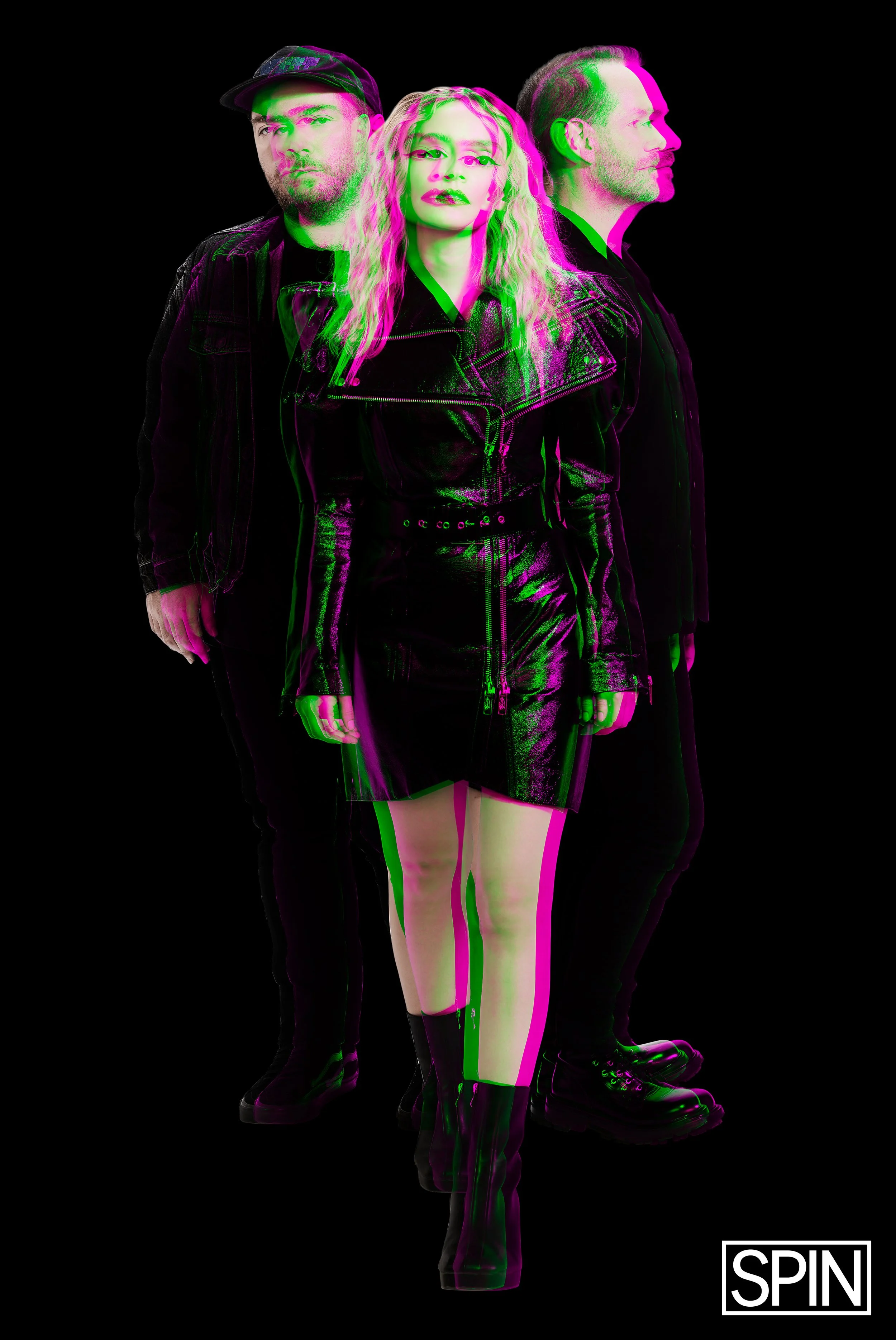 Three people standing side by side, with a visual glitch effect, against a black background. The person in the center is a woman with wavy blonde hair, wearing a black leather jacket, skirt, and boots. The two men on either side are dressed in black 