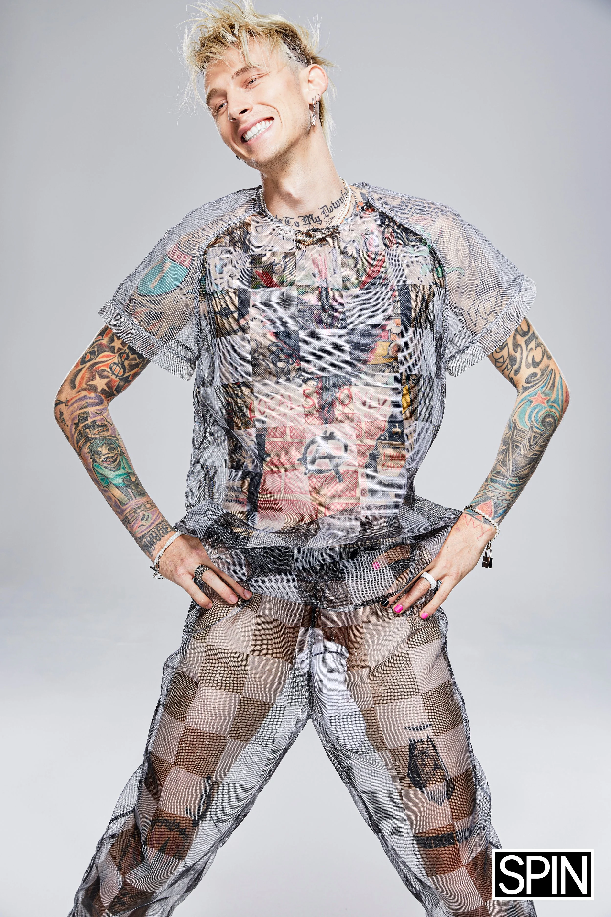 A young man with blonde hair and multiple tattoos posing in a confident stance, wearing a sheer, graphic t-shirt and checkered sheer pants, smiling with his eyes closed.