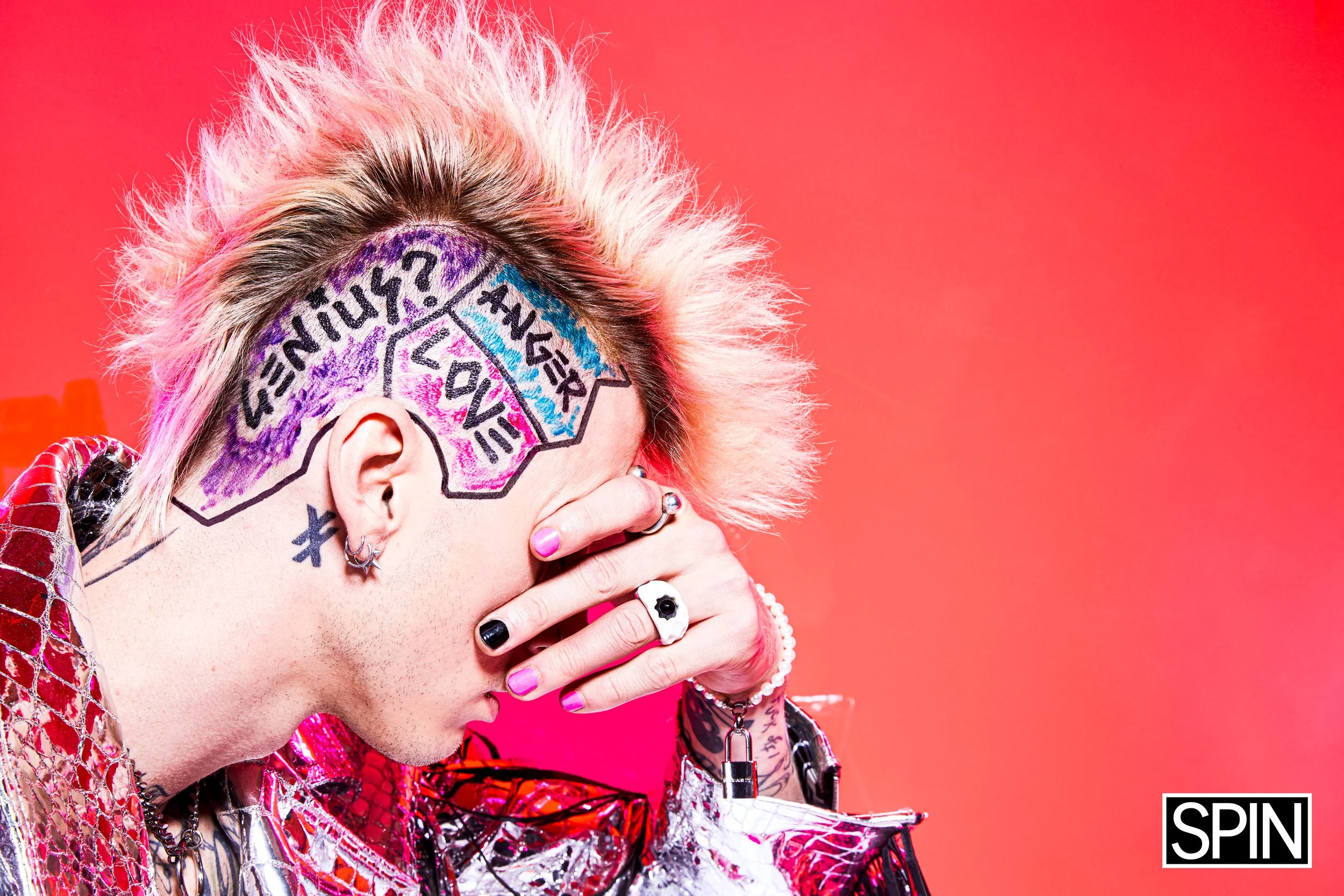 A person with a colorful mohawk hairstyle and tattoos, covering their face with their hand, stands against a pink background. They are wearing a shiny, patterned jacket and various jewelry.
