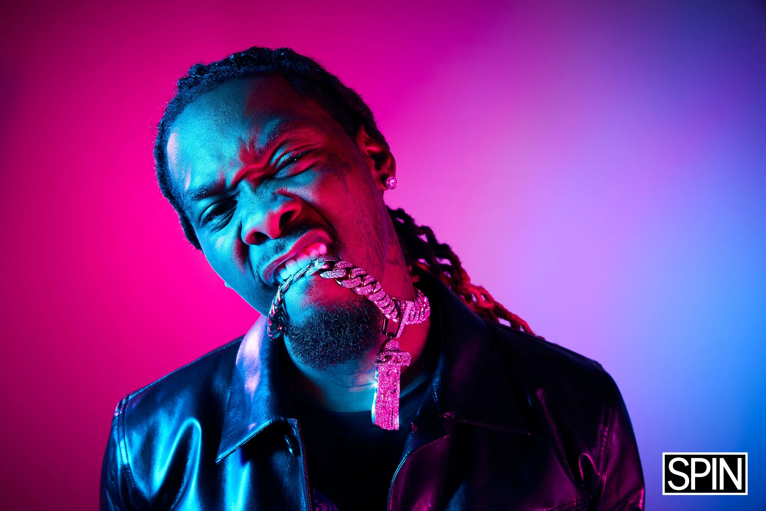 A man with long hair and a beard showing his teeth with a gold grill, wearing a leather jacket and multiple jewelry including a chain with a cross pendant, against a pink and purple gradient background.