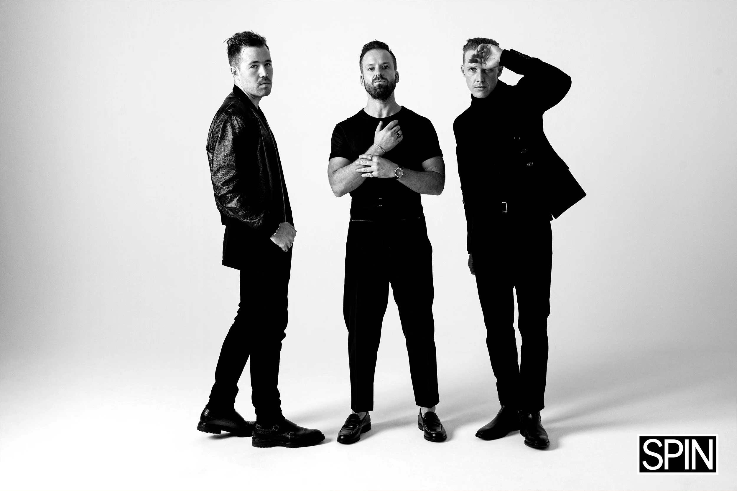 Black and white photo of three men standing side by side against a plain background. The man on the left is wearing a leather jacket, the man in the middle is in a t-shirt with hands on his chest, and the man on the right is in a fitted coat with one