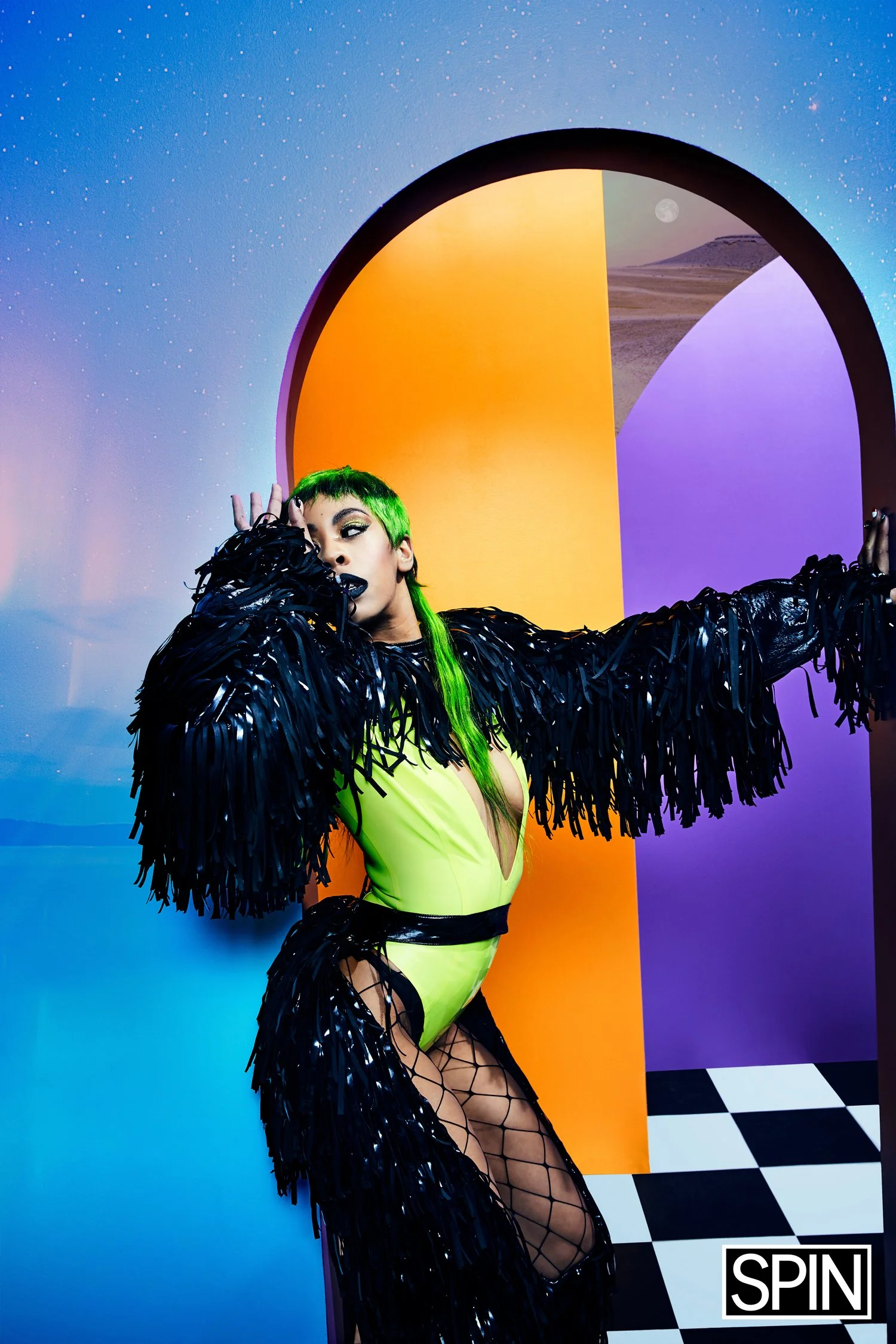 A woman with bright green hair wearing a neon yellow and black outfit with black fringed sleeves, posing in front of a colorful, abstract background with a checkered floor and the word 'SPIN' in the bottom corner.
