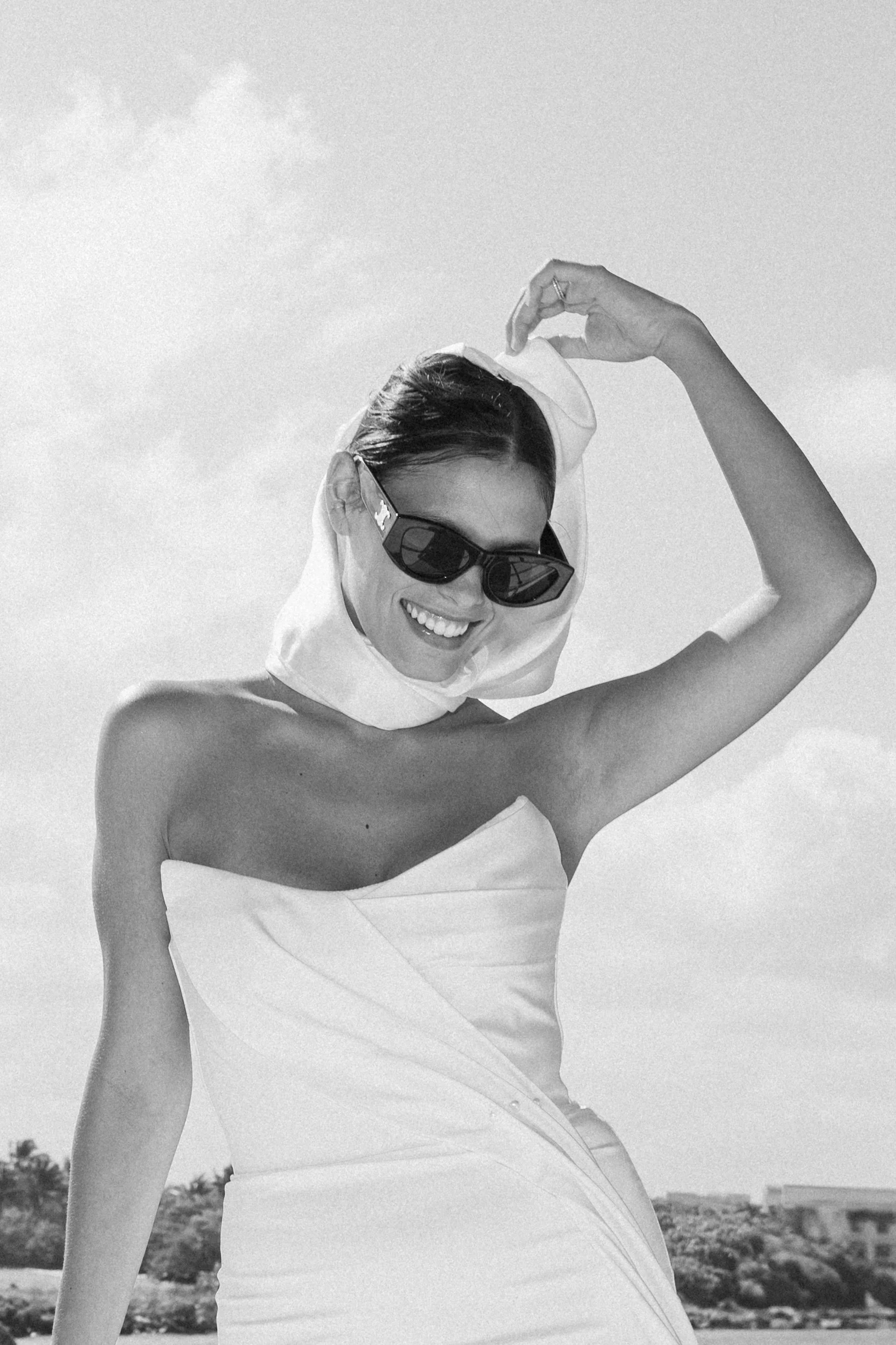 A woman smiling and wearing sunglasses, holding a cloth to her head, wearing a strapless dress, outdoors with trees and buildings in the background.