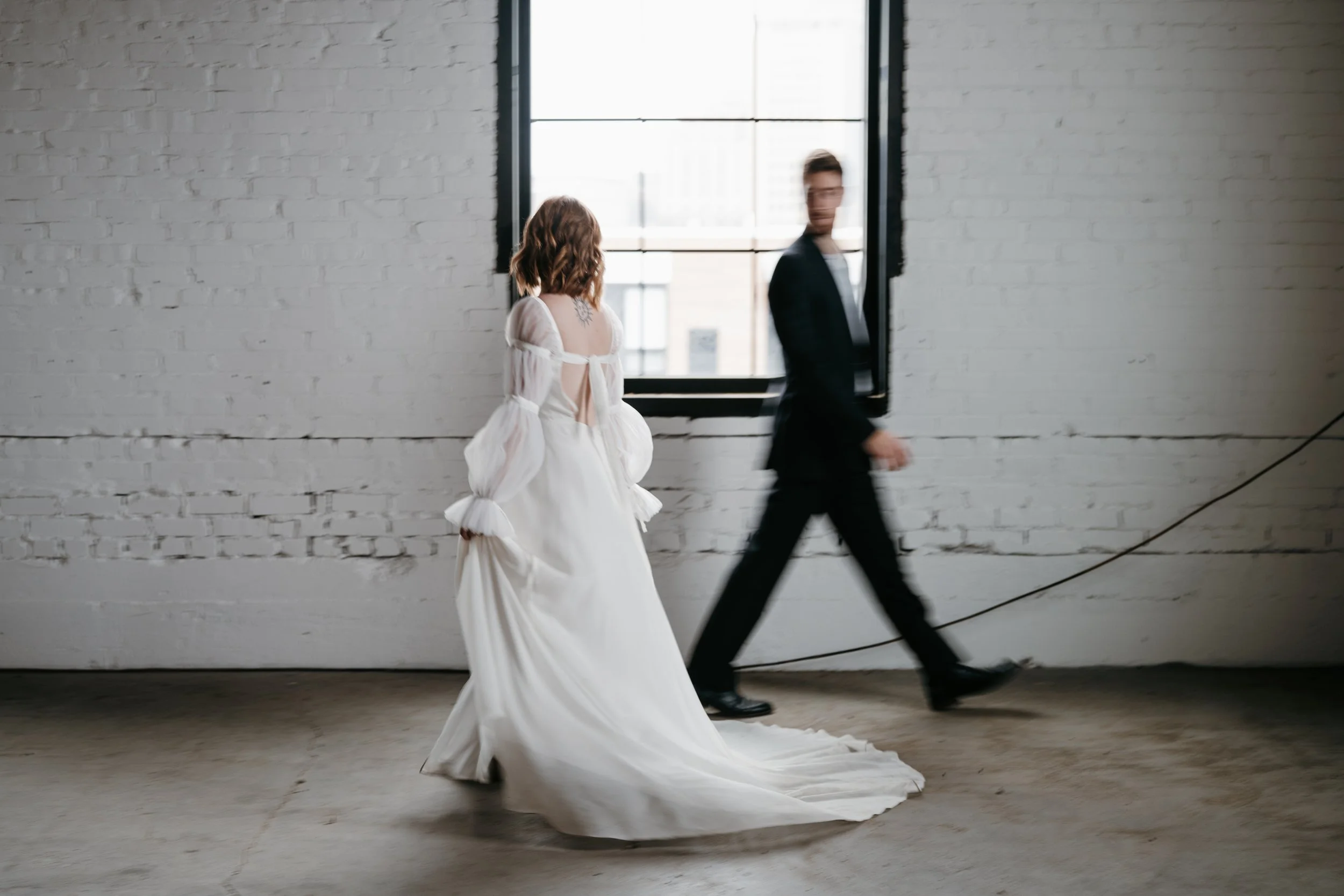 A woman in a white wedding dress walking in front of a man in a black suit in an industrial-style room with white brick walls and a large window.