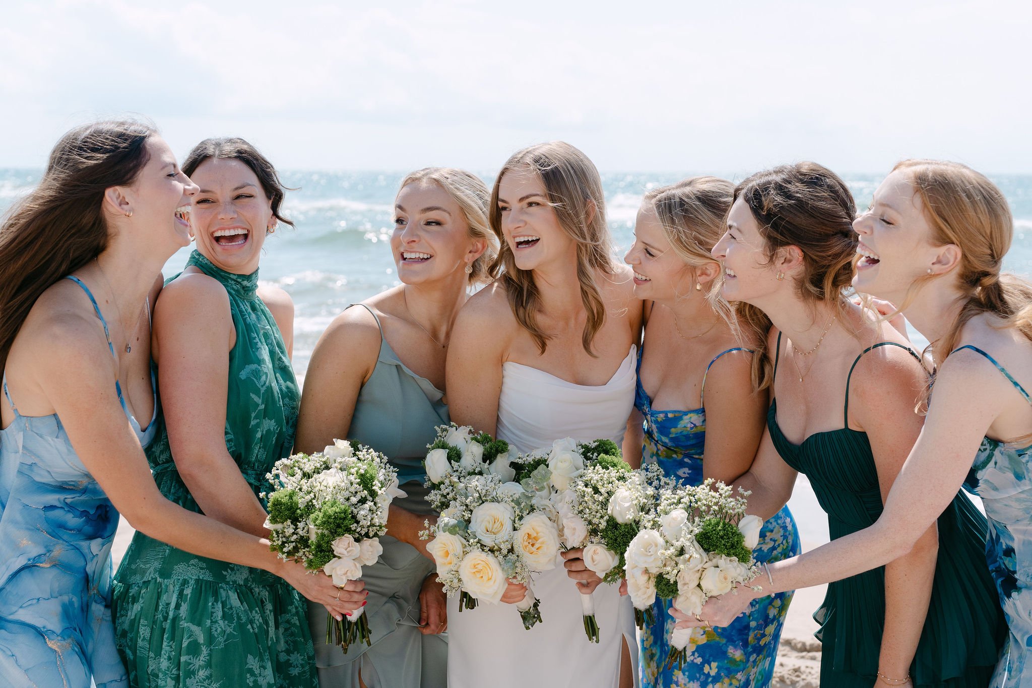 Group of women dressed in summer dresses standing on the beach holding bouquets of flowers, smiling and laughing.