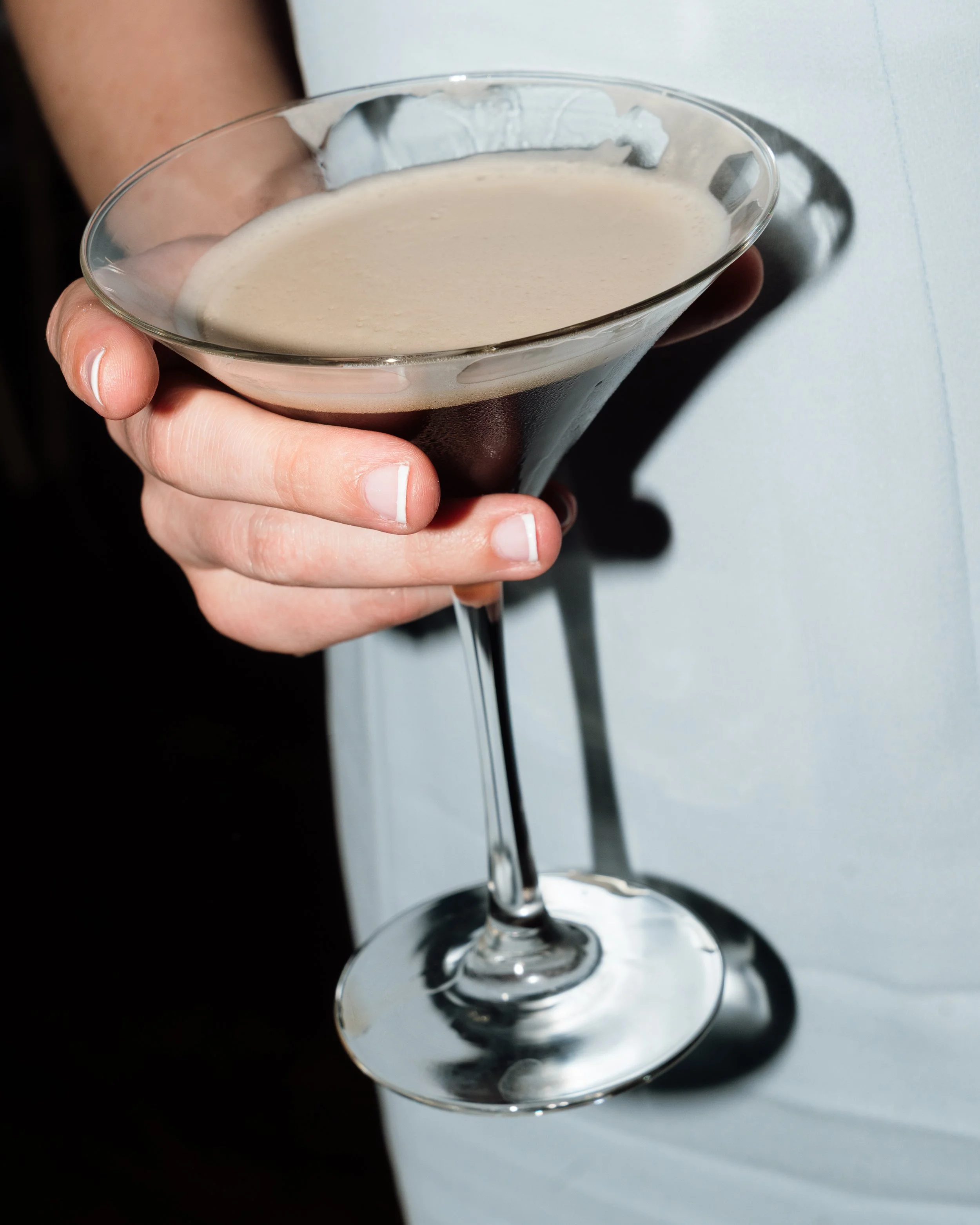 A person's hand holding a martini glass filled with a creamy, beige-colored cocktail, against a plain light background.