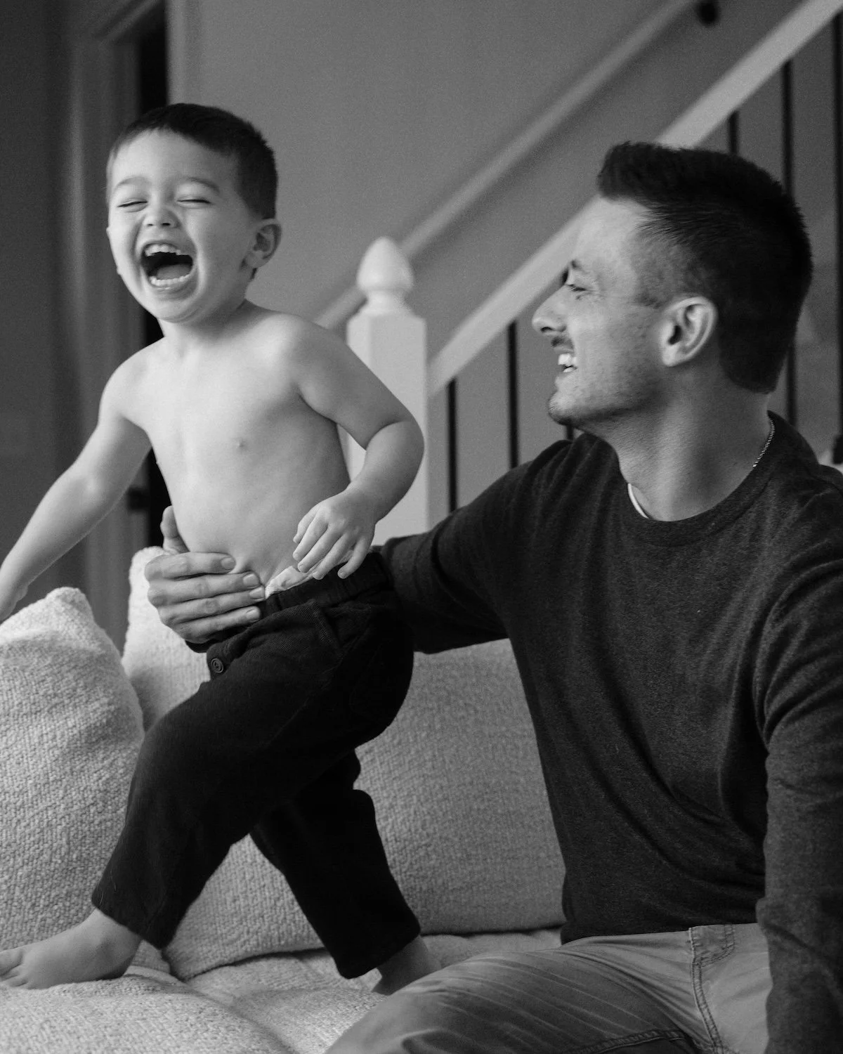 Dad is holding son on couch, son is laughing, trying to run away from dad