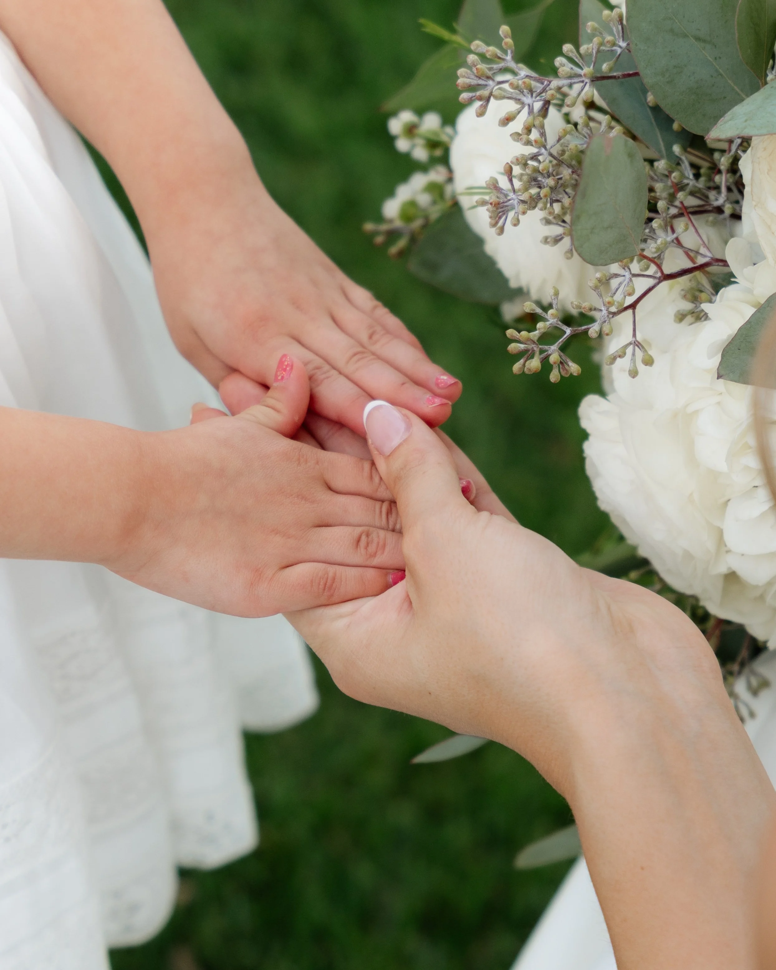 Two hands, one adult and one child, gently holding each other near a white floral arrangement outdoors.