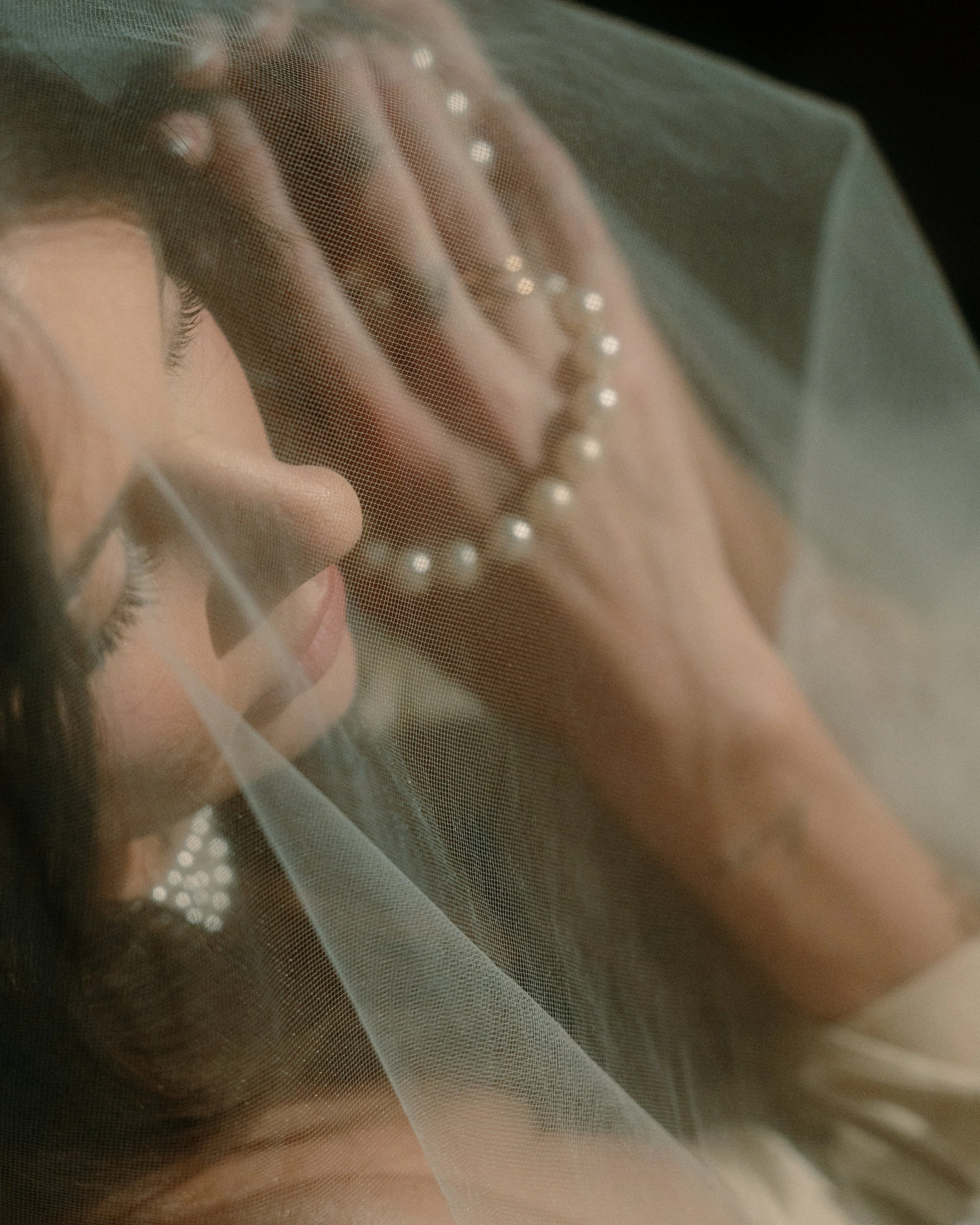 A woman with closed eyes, wearing a veil and a pearl necklace, viewed through a sheer fabric.