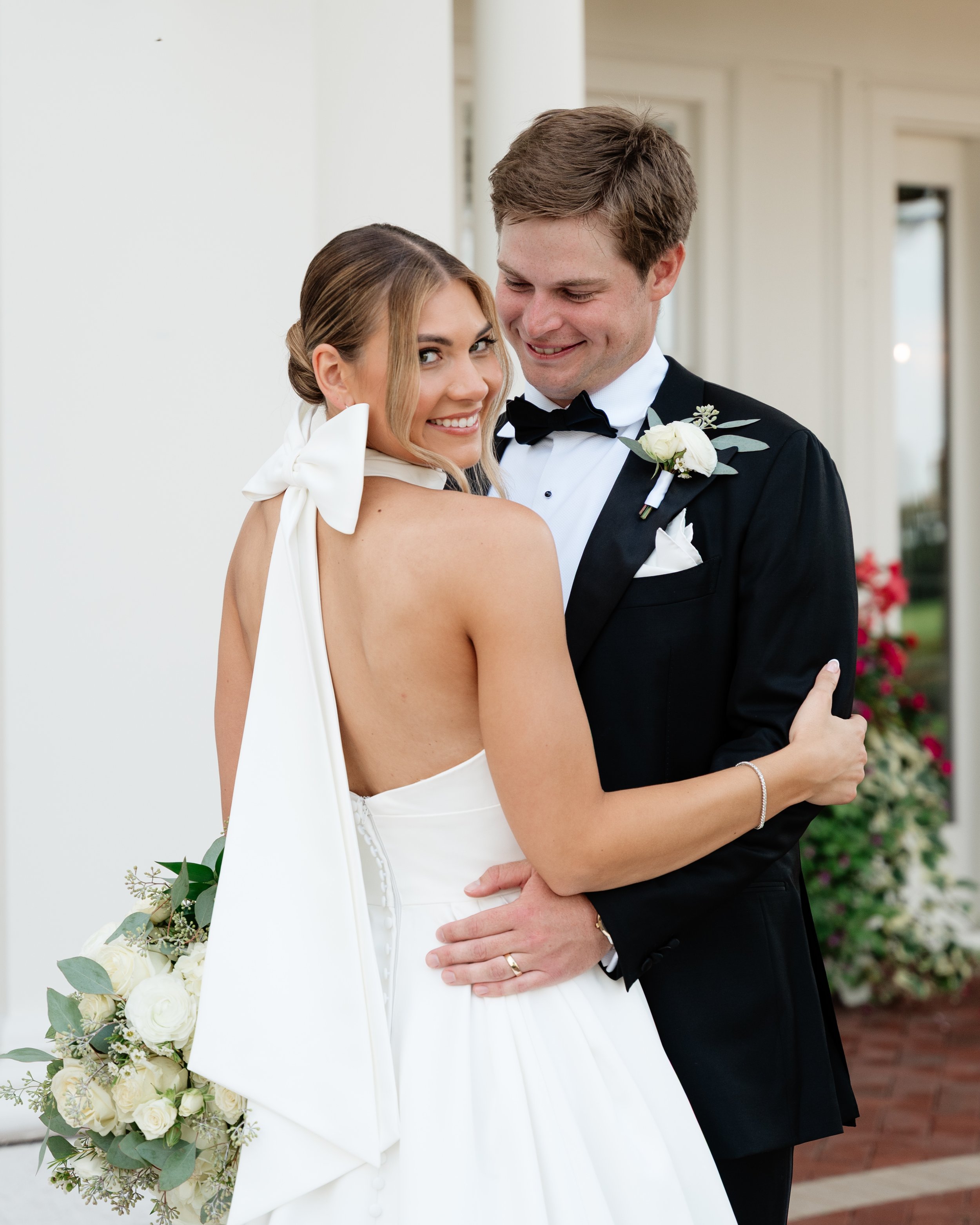 A newlywed couple in wedding attire smiling and embracing outside a building with white walls and flowers.