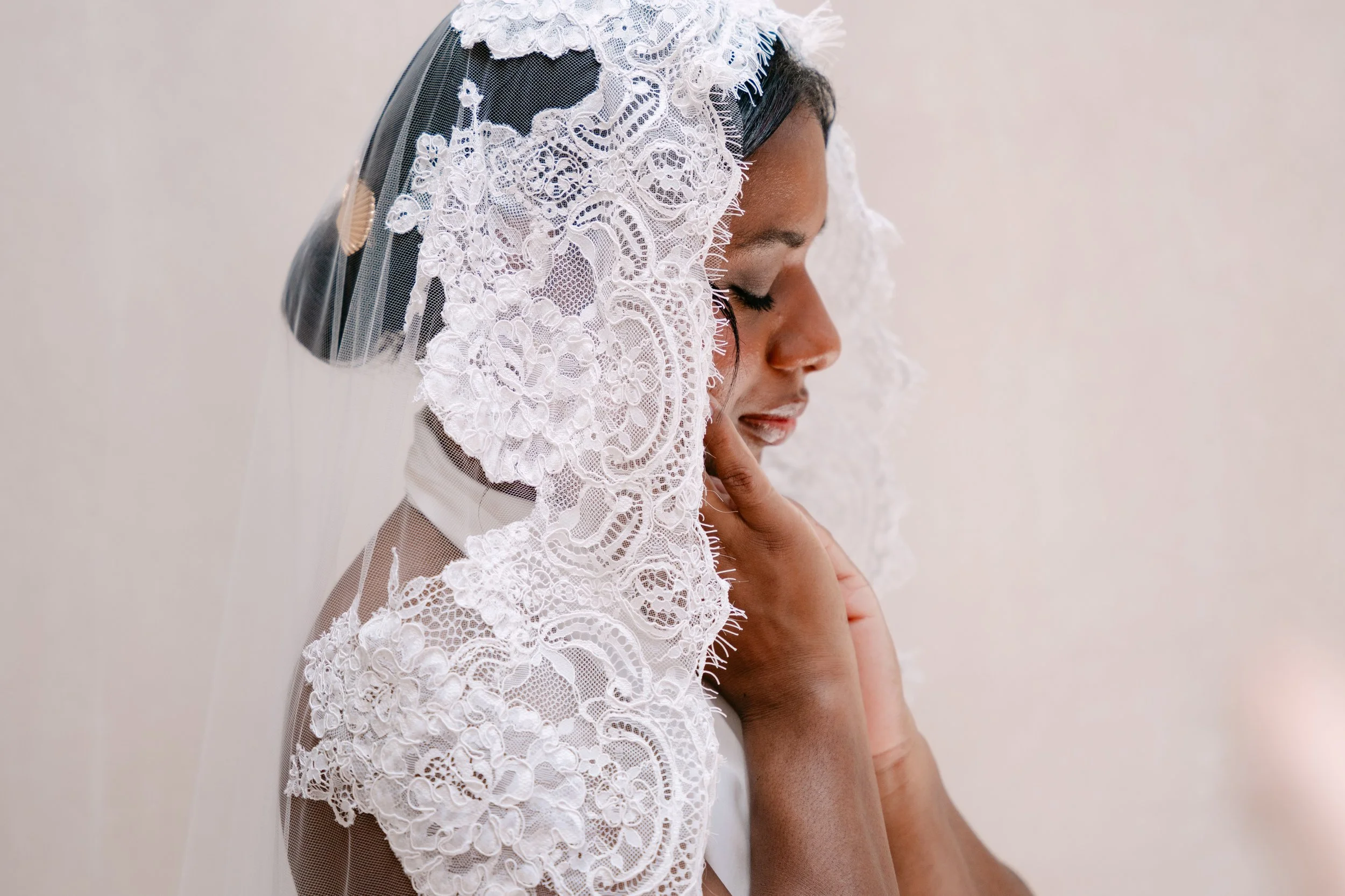 A woman with dark hair, closed eyes, and a peaceful expression wearing a lace veil with floral patterns, touching her face gently.
