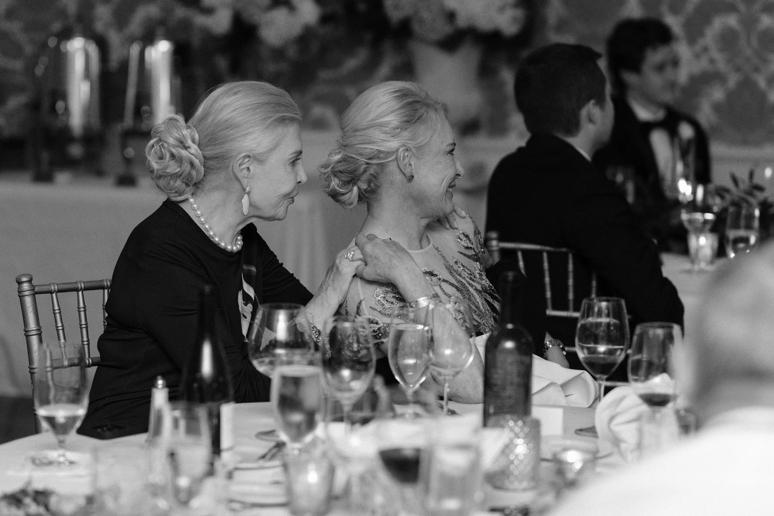 Women and men dressed formally are seated at a banquet table in an upscale dining setting, smiling and engaging in conversation.