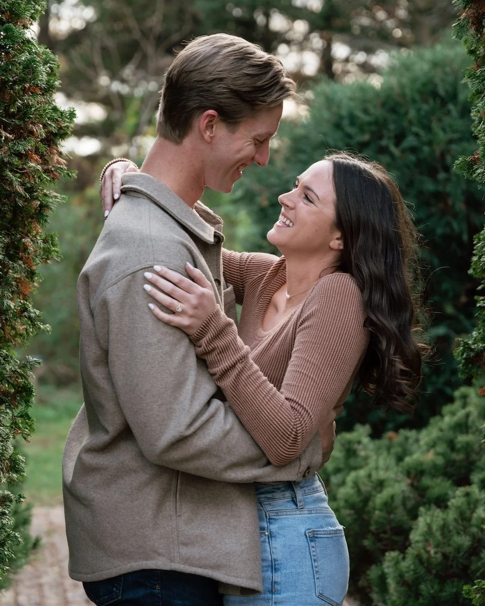A young couple embracing and smiling at each other outdoors among green trees.