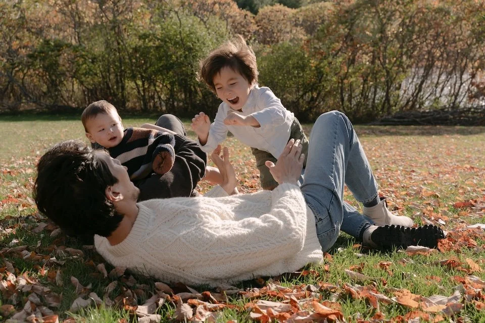 family photo session, young boy is midair, jumping to dad who is lying down on the grass, fall leaves surround the family.