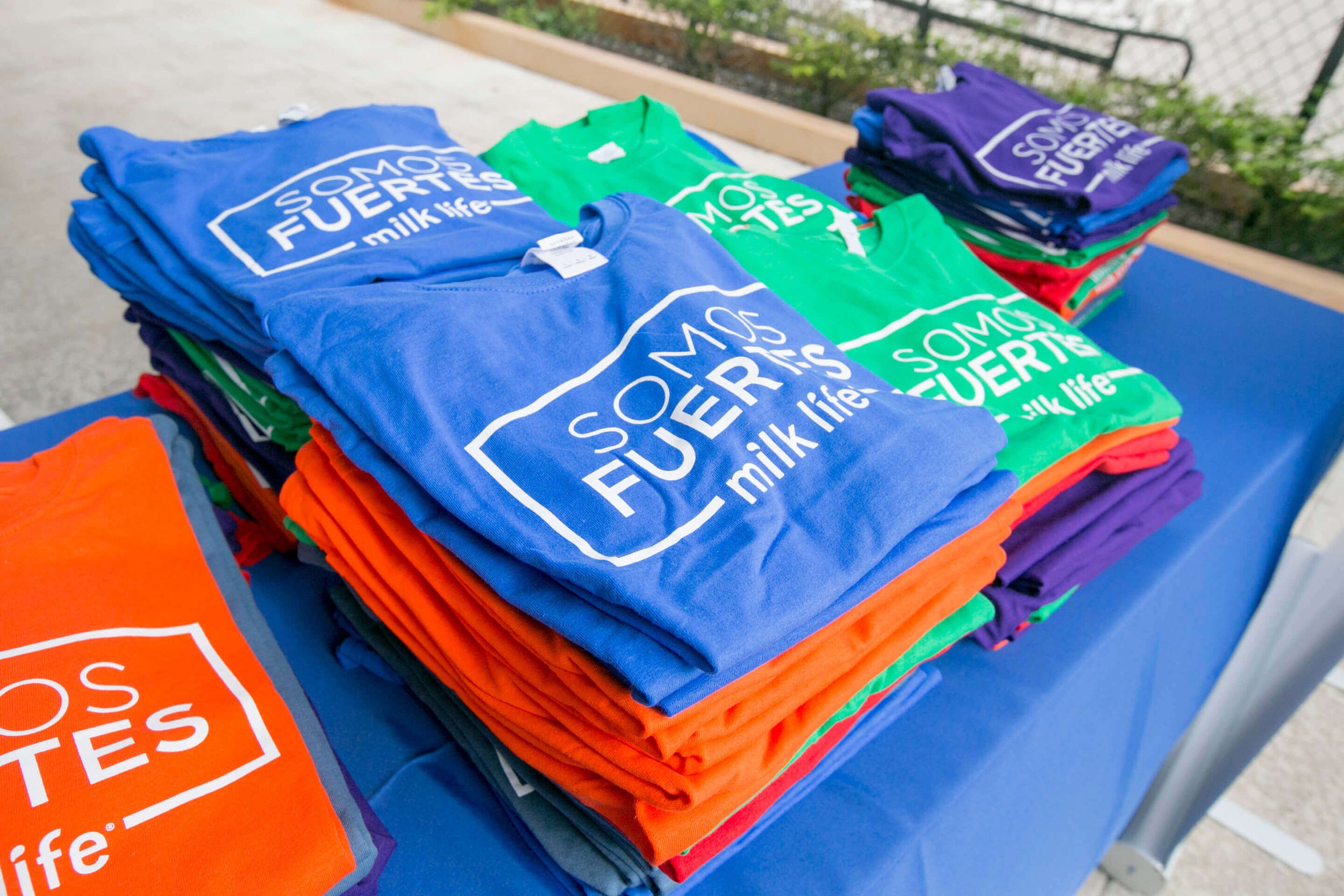 Event t-shirts in blue, orange, green, and purple that say "Somos Fuertes, Milk Life"