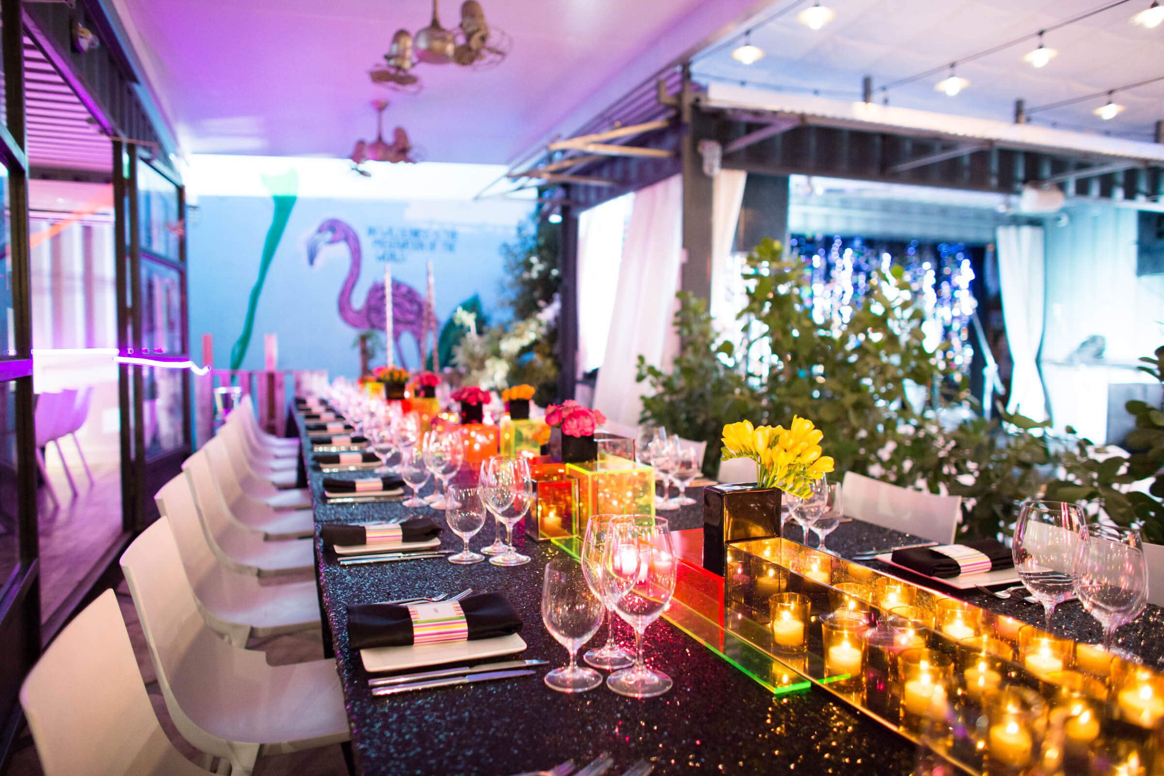 Modern dinner party set up on a black table full of candles and neon. A Flamingo on the wall in the background.