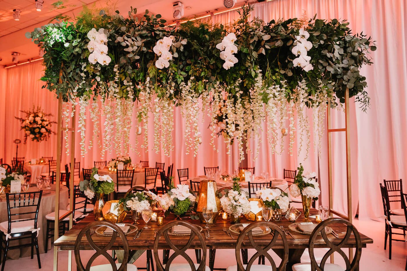 Wooden vineyard wedding table set under a leafy hanging floral in front of white pipe and drape with pink lighting.