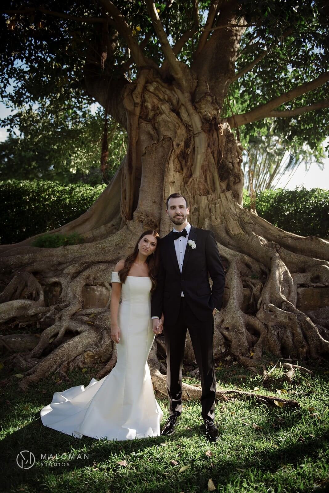 Bride and groom, smiling, stare directly into camera under a tree