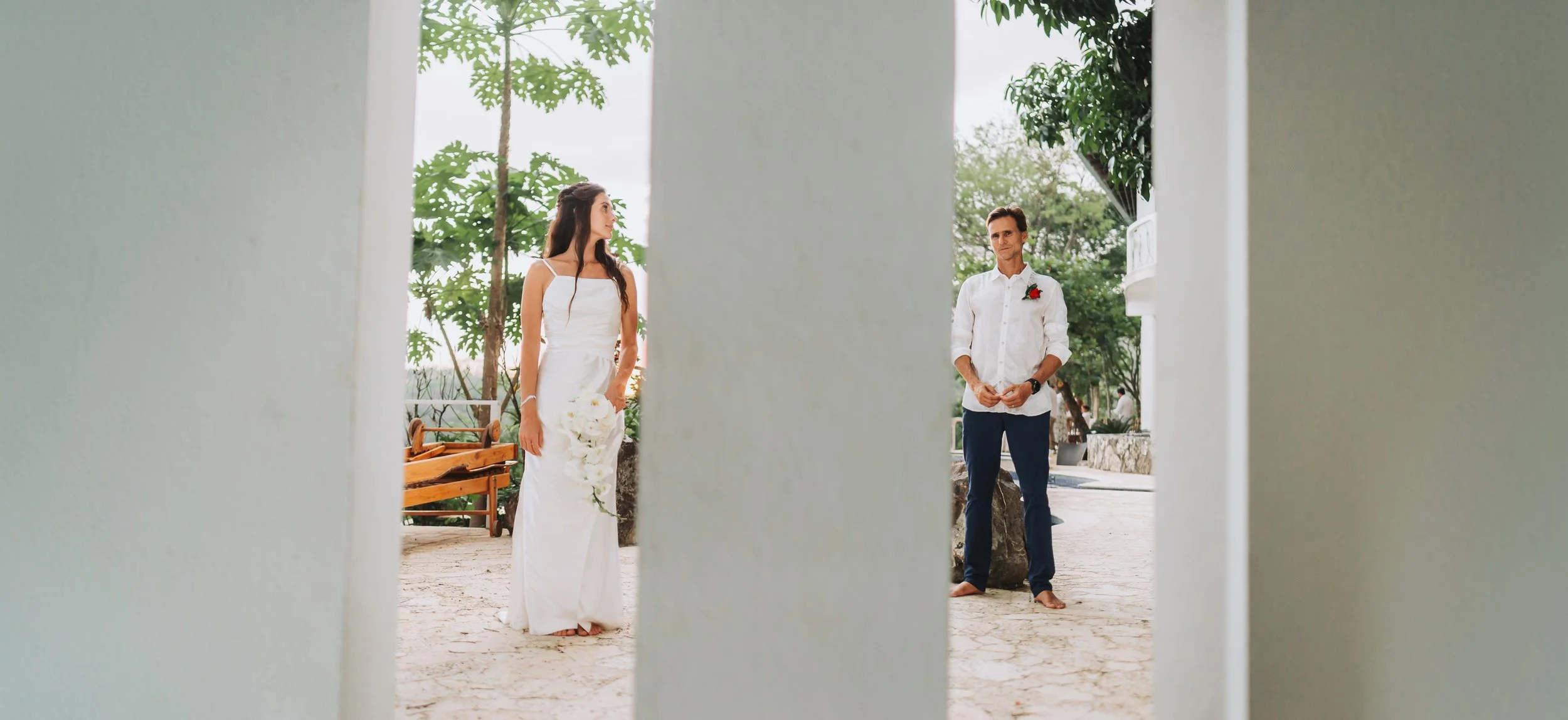 A woman in a white wedding dress holding a bouquet of white flowers and a man in a white shirt with rolled-up sleeves, dark pants, and a red boutonniere, standing outdoors on a stone-paved area with trees and a building in the background, seen throug